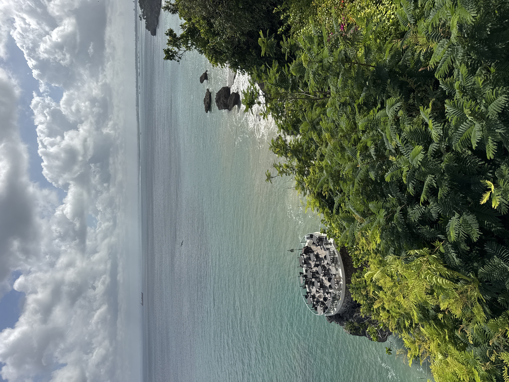 The image shows a coastal view in Kuta Selatan, Indonesia, featuring a circular clifftop bar or restaurant. The establishment, equipped with tables and chairs, juts out over the ocean. Lush, green vegetation covers the adjacent cliffside. The sea appears calm and turquoise in color, extending to a horizon under a cloudy sky. A ship is visible on the horizon, and a bird is in flight. The photograph is taken from an elevated perspective, looking down at the scene.