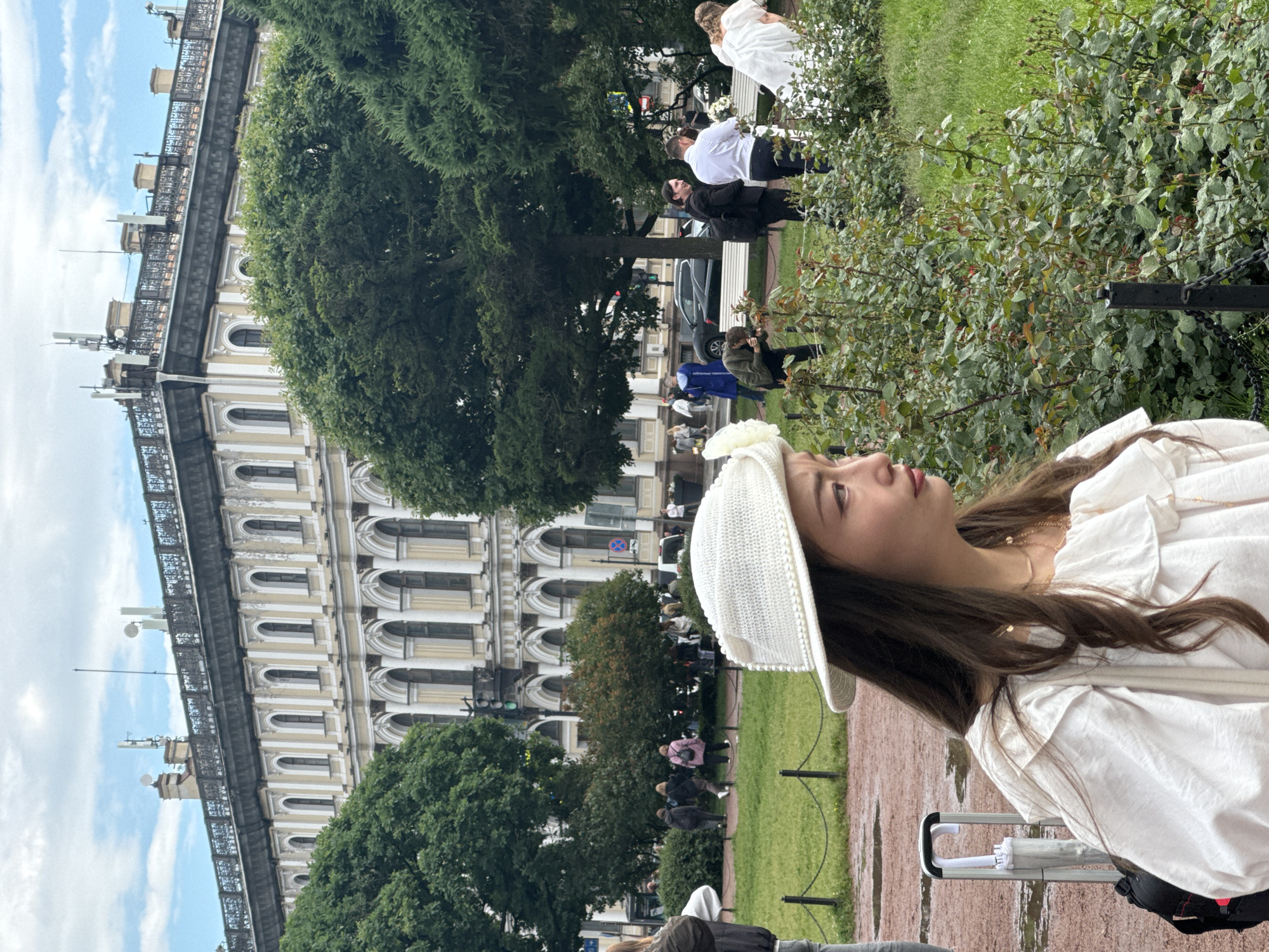 The image depicts a group of people gathered in a park-like setting with a grand, ornate building in the background. The architecture of the building, with its arched windows and classical detailing, suggests it is a historic structure in Saint Petersburg, Russia. In the foreground, a woman wearing a white hat and a white ruffled dress is looking to the right, partially obscuring a bouquet of flowers on her right. Behind her, to the right, a wedding party appears to be in session, with a bride in a white gown and several other individuals in formal attire. Some are engaged in conversation, while one person is taking photographs. The environment is characterized by green trees and manicured bushes, with a paved walkway and some patches of wet ground, indicating recent rain. The sky is cloudy with patches of blue.