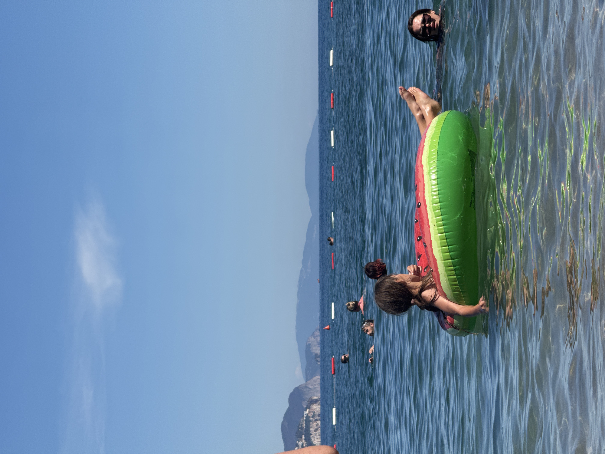 A bright, sunny day at a beach in Budva, Montenegro. The clear blue sky is mostly cloudless, with a faint wispy cloud in the distance. The sea is a deep blue, with gentle waves reflecting the sunlight. In the foreground, a young girl with dark hair tied in a ponytail is floating on her back in a watermelon-shaped inflatable ring. Her legs are extended, and she appears relaxed. To her right, a woman with dark hair and sunglasses is partially submerged in the water, looking towards the camera. Further out in the water, several other people are visible, swimming and enjoying the sea. In the background, a line of buoys marks the swimming area, and beyond that, the coastline is lined with hills and buildings. The overall scene conveys a sense of summer vacation and relaxation.