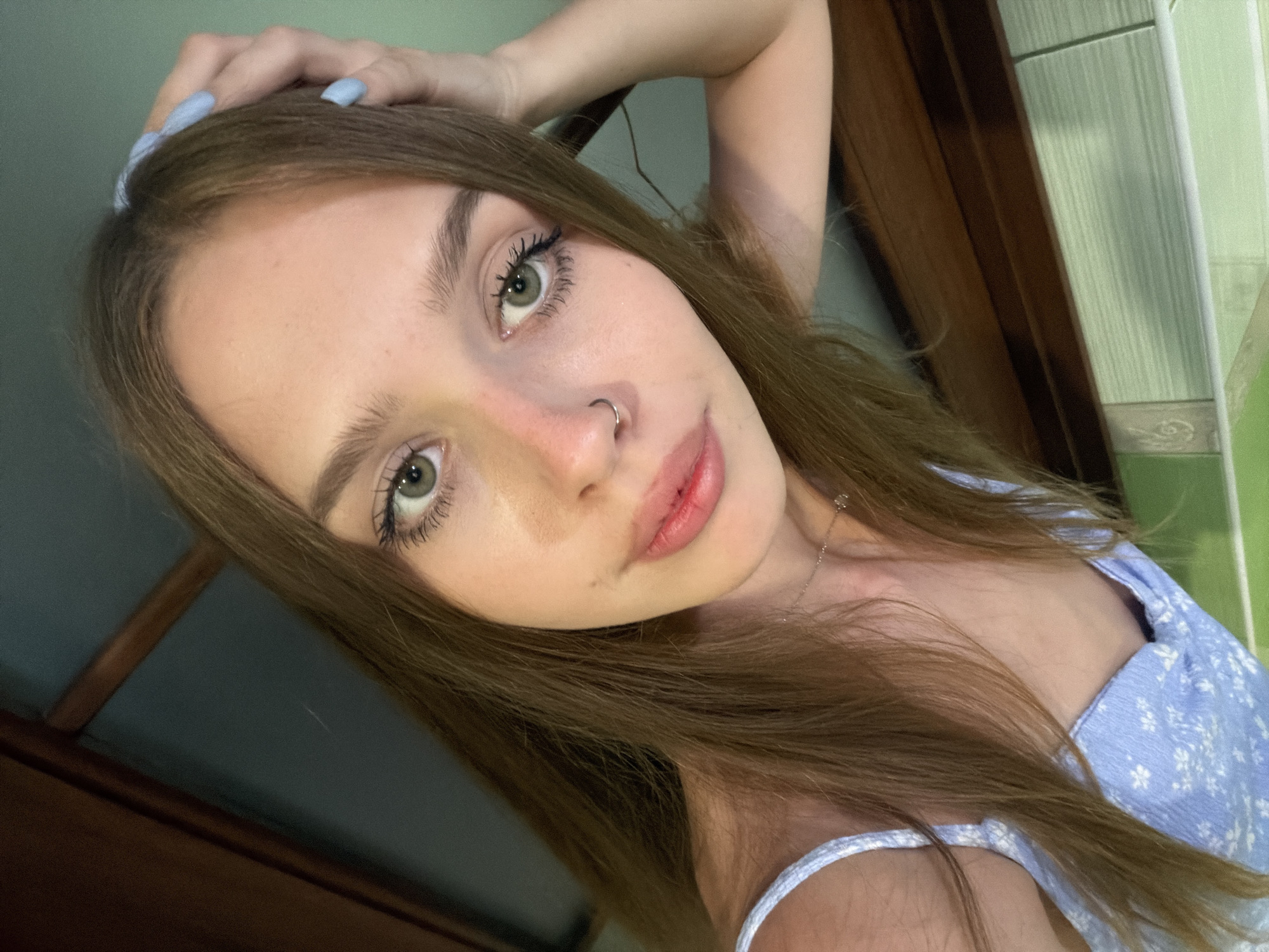 A young woman with long, brown hair and striking green eyes is captured in a close-up, tilted selfie. Her hand, with pale blue-painted fingernails, is resting on the top of her head. She is wearing a light blue floral dress with thin straps. A delicate silver necklace is visible around her neck, and she has a small silver hoop in her nose. Her lips are slightly parted, and she appears to be looking directly at the camera with a neutral or slightly playful expression. The background is softly blurred, showing hints of a wooden surface and a green, tiled wall, suggesting an indoor setting. The lighting is relatively soft, creating a gentle glow on her skin. The image does not contain any visible text. The overall impression is that of a casual, personal snapshot, possibly taken indoors during the daytime.