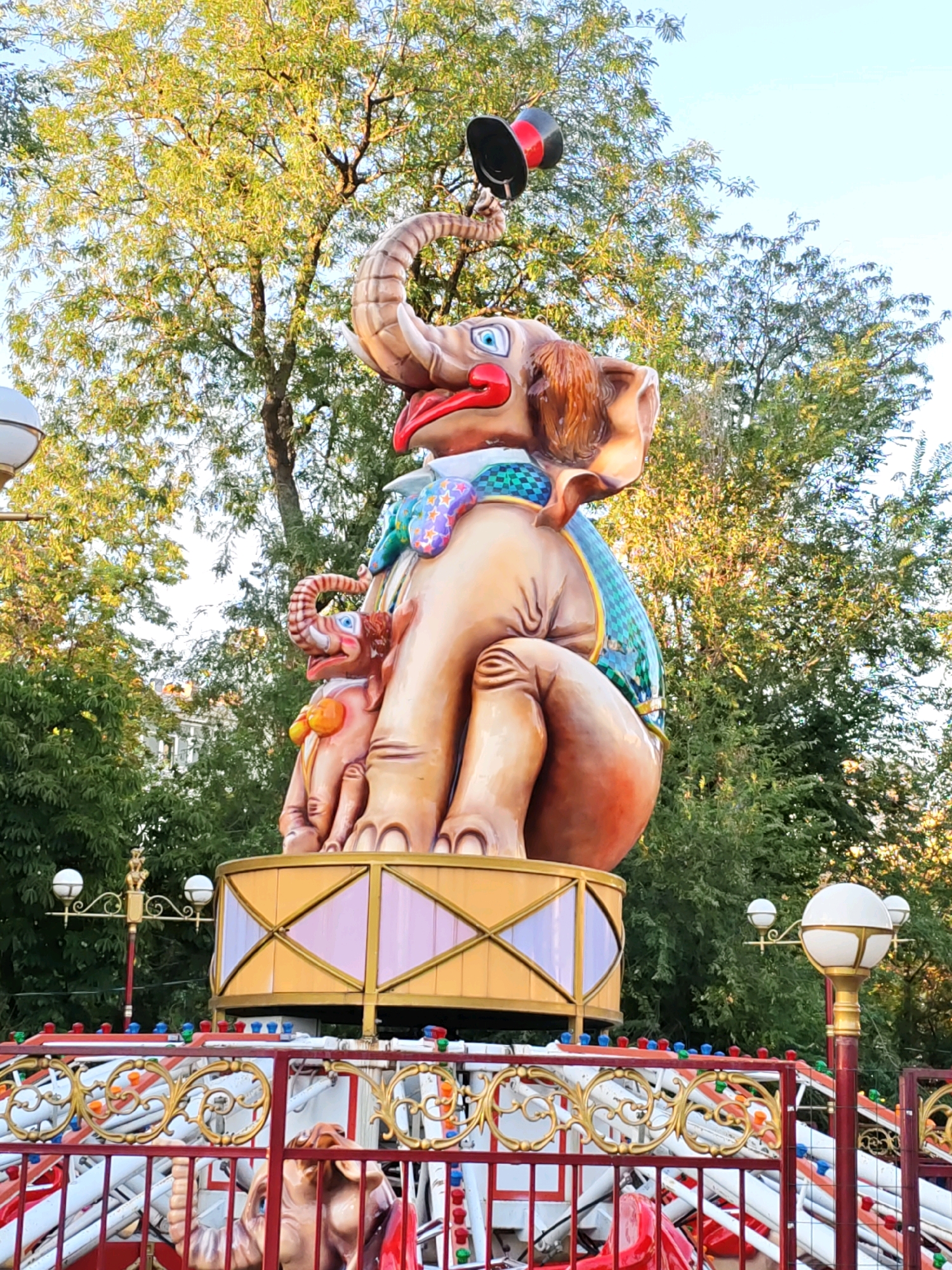 A large, colorful, and whimsical statue of two elephants, one adult and one baby, is the central focus of this outdoor scene, likely an amusement park or fairground attraction in Rostov-on-Don, Russia. The adult elephant, positioned on a raised platform, is depicted with its trunk raised, balancing a black top hat with a red band. It wears a vibrant, patterned bib and a green and blue checkered blanket. The baby elephant sits nestled beside the adult, also looking upward. Both elephants have blue eyes and red lips. The platform they stand on is adorned with decorative gold trim and panels featuring pink and yellow geometric shapes.
In the foreground, a red and gold railing of another ride is visible, with colorful lights strung along its top. The environment is lush with green trees and foliage in the background, suggesting a warm, sunny day. The sky is a clear light blue. Streetlights with white spherical globes are also present, one with a golden decorative element atop its pole, another more prominent one to the right. The overall impression is one of cheerful, playful decoration characteristic of a public entertainment venue.