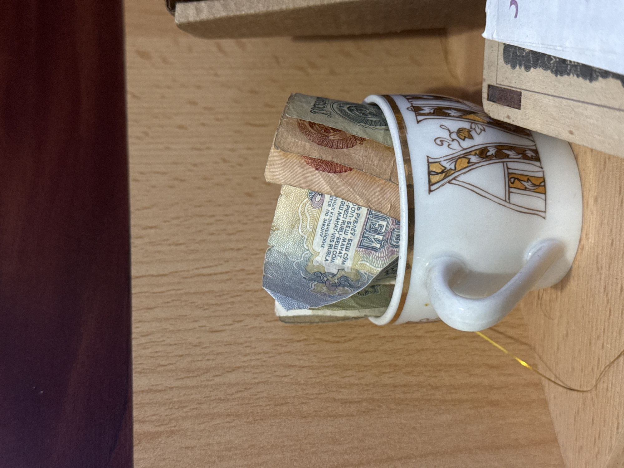 The image displays a collection of old banknotes stored within a white teacup, resting on a light-colored wooden surface in Kladovitsy, Russia. The teacup, positioned horizontally, features a decorative pattern in gold and brown, possibly stylized foliage or an ornate letter, and has a handle.
Several banknotes are visible, folded and tucked inside the cup. They appear aged and worn, with colors predominantly in muted greens, browns, and blues. Text on the notes, confirmed by OCR, includes phrases in multiple languages, indicative of Soviet-era currency. Specifically, fragments like "ДЬ РУБЛЁЎ БЕШ СЎМ," "БОЛ БЕШ МАНАТ," "PIECI RUBĻI БЕШ СОМ," and "SƏLI MAHAT VIIS RUBLA" are discernible, all translating to variations of "five rubles" or local currency units. Additional text such as "ННЫХ КАЗНАЧЕЙСКИХ" (of treasury) and "ЕТСЯ ПО ЗАКОНУ" (according to the law) is also present. Some banknotes exhibit what appears to be the emblem of the Soviet Union.
In the background, a dark reddish-brown vertical surface is visible on the left, and a lighter, cardboard-like object is partially seen above the cup. A thin, golden string or wire lies near the cup's handle on the wooden surface. The scene captures a nostalgic arrangement, possibly a memento or a numismatic collection.