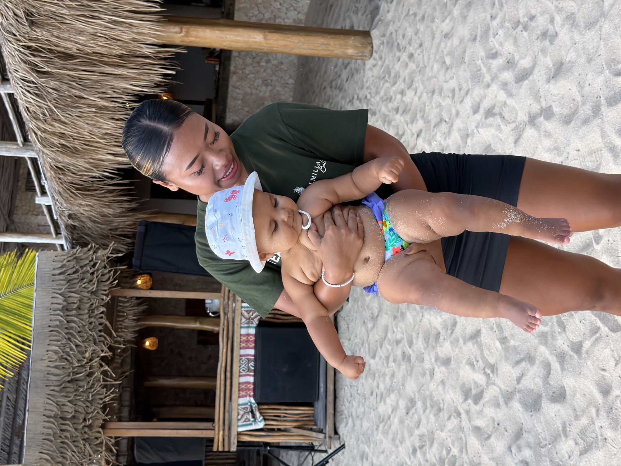 A woman holds a baby on a sandy beach in Kuta Selatan, Indonesia. The woman is wearing a dark green t-shirt and black shorts, and her hair is pulled back in a bun with highlights. She is smiling down at the baby, who is wearing a white hat with colorful patterns and a swim diaper with a floral design. The baby has a pacifier in its mouth and a white bracelet on its wrist. The scene appears to be outdoors during the day, with sandy ground and thatched-roof structures in the background. There is also some greenery visible, possibly palm leaves. The overall mood seems relaxed and happy, suggesting a casual day at the beach. A portion of text is visible on the woman's t-shirt, though it is partially obscured and difficult to read.