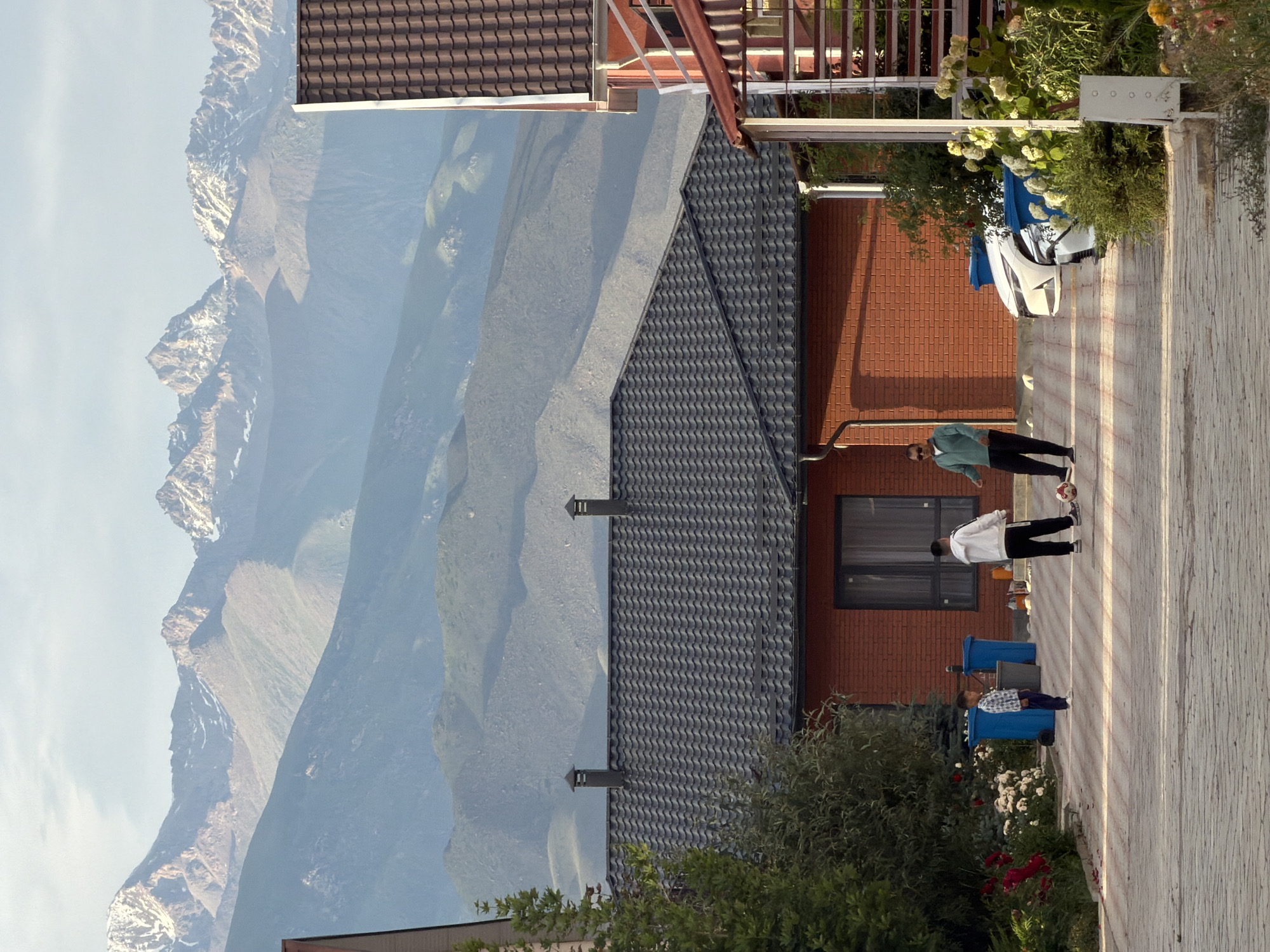 This is a vertically oriented, eye-level shot of a residential scene set against a backdrop of majestic mountains. The foreground features a paved walkway leading past a brick house with a dark, textured roof and a prominent chimney. Two people are visible on the walkway. One, wearing a light green jacket and dark pants, stands facing away from the camera. Another, dressed in a white hooded jacket and dark pants, is bending down, perhaps interacting with something on the ground. To the left of the walkway, a white car is partially visible, covered by a blue tarp. Beyond the house, the terrain slopes upwards, revealing the layered geological formations of distant, rugged mountains. These mountains are rendered in muted blues and grays, with some patches of snow visible on their upper reaches, suggesting a cool climate. The sky is a pale, hazy blue, indicating a clear day. The lighting suggests it's either late afternoon or early morning, casting long shadows on the walkway. The overall impression is one of a serene, possibly remote, dwelling nestled in a dramatic natural landscape.