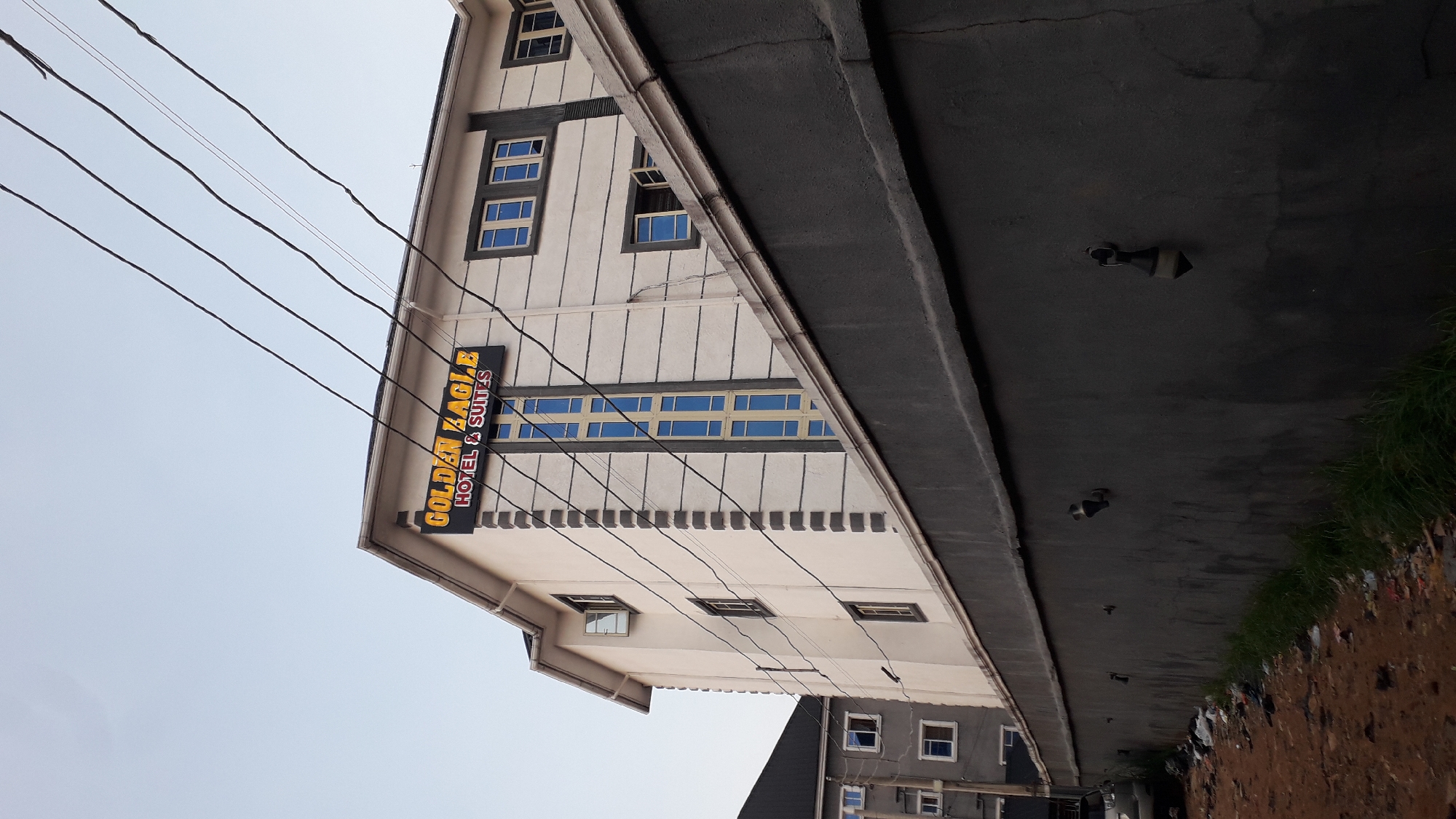 The image depicts the "Golden Eagle Hotel & Suites" in Nkpor, Nigeria, captured from an unconventional low-angle perspective. The building has a triangular facade. Above it is the sky and a tangle of electrical wires are visible. Along the top right, an overpass or elevated road appears, with mounted lights along its underside and a patch of green growth bordering the ground below. In the distance, a section of another building with windows is visible.