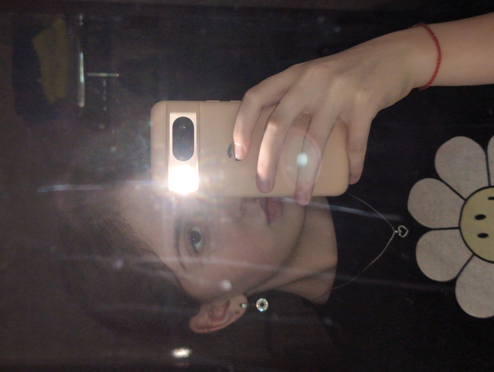 A person takes a selfie in a mirror with their phone. The phone's flash is on, obscuring some facial features. The person is wearing a dark-colored shirt with a large white flower design. They have an earring in their left ear and a red bracelet on their left wrist. A necklace with a heart pendant is visible. The location is Unknown, Unknown.