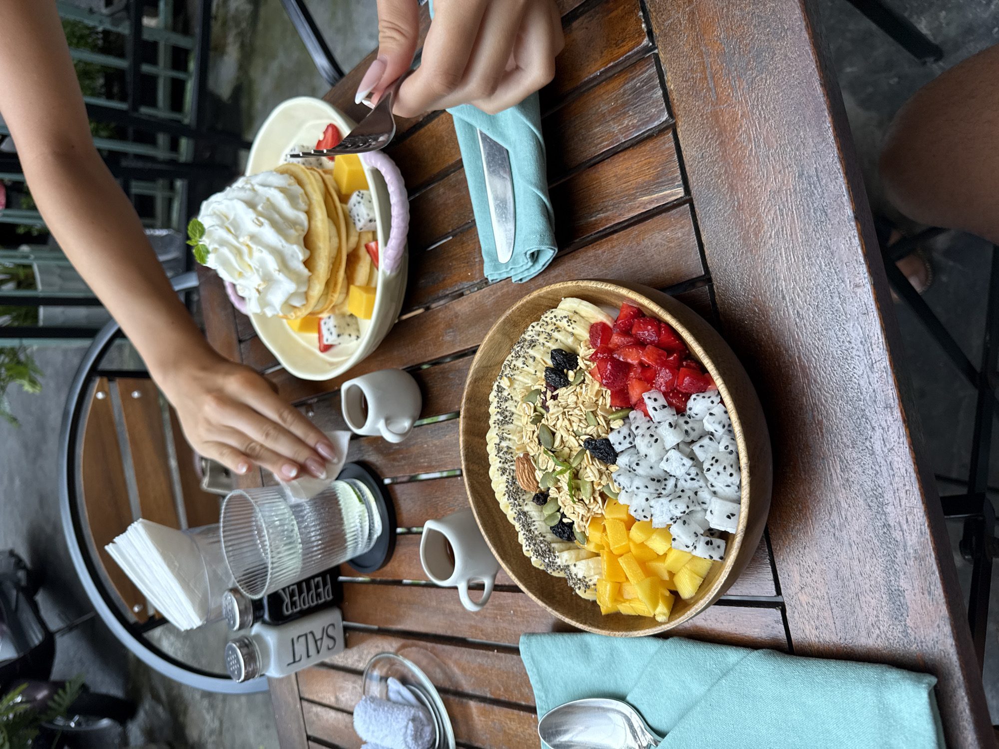 The image shows a wooden table at a restaurant in Đà Nẵng, Vietnam. Two people are visible, each with a plate of food. One person is about to eat pancakes topped with whipped cream, mango, and dragon fruit. The other has a fruit bowl with banana, strawberries, dragon fruit, mango, and granola. A fork is in the person's hand with the pancakes. A glass of water, salt, pepper, and napkins are on a small table next to the person with the pancakes. The weather appears to be overcast.