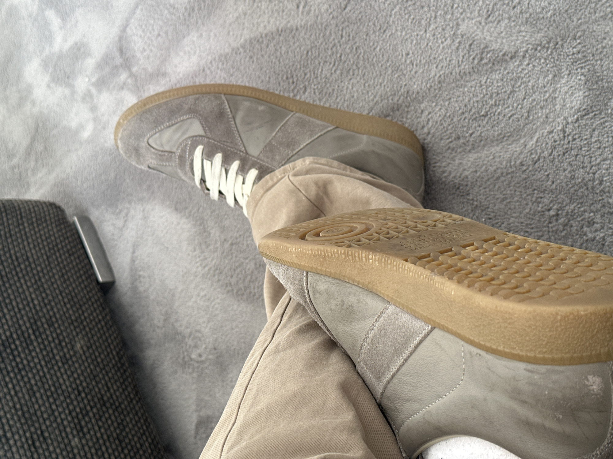 The image shows a person sitting with their legs crossed, wearing gray sneakers with white laces and tan pants. The sneakers appear to be overlapping. The background consists of a gray, textured surface. A portion of a gray upholstered chair is visible on the left side of the image. The location and the city of Milan, Italy, are not evident from the visual information provided.