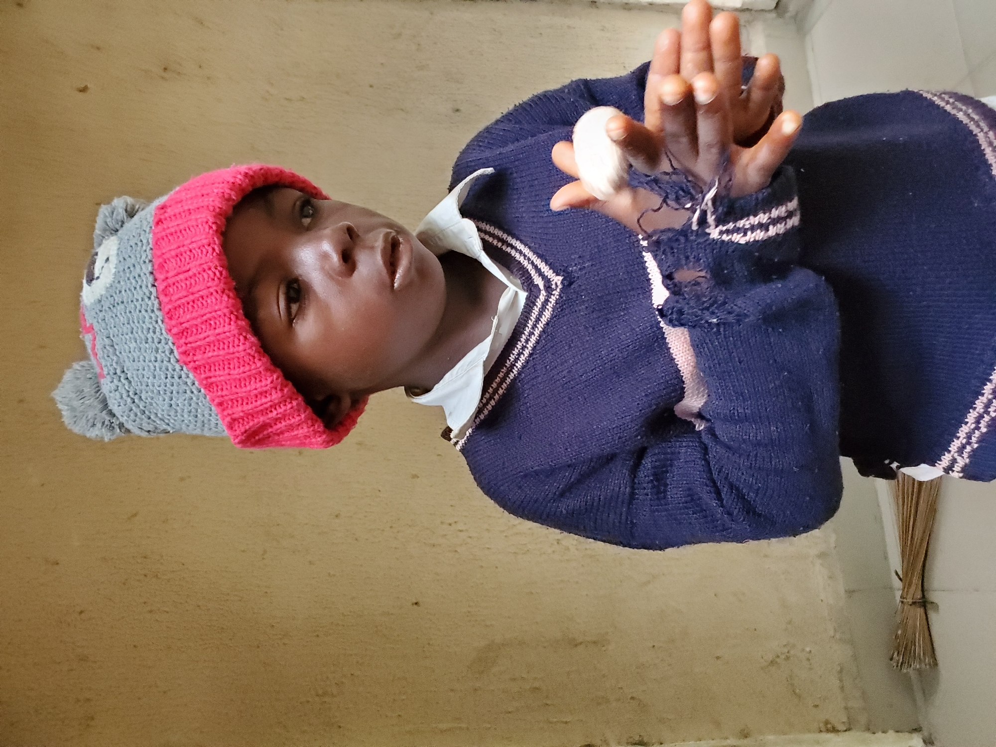 A child wearing a gray and pink knit hat and a navy blue sweater stands indoors. The sweater has a white collar and a V-neck with white trim. The child is holding a white object in their hands. The wrist of the sweater is frayed and torn. The background includes a beige wall, and a bundle of sticks tied together is leaning against a white surface. The location is Mararaba, Nigeria.