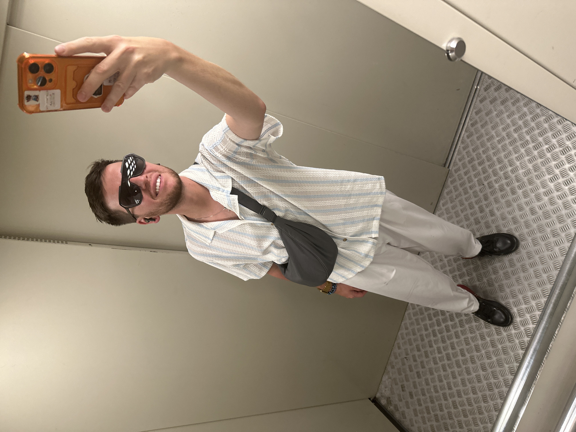 Here's a detailed description of the image:
**Scene:** The image appears to be a selfie taken by a person in what seems to be an elevator or a small service area.
**Person:** A person is visible in the photo, facing the reflection in a mirror. They have short brown hair and a beard. They are wearing sunglasses, a striped shirt, light-colored pants, and black shoes. A gray bag is slung across their body. They are holding a smartphone with an orange case, presumably taking the picture. They are smiling.
**Environment:** The background consists of plain walls. The floor is covered with a metallic tread plate, commonly found in industrial settings or elevators. There is a silver button visible near the corner of the elevator car.
**Action:** The person is taking a selfie.
**Objects:**
* Smartphone with an orange case
* Sunglasses
* Striped shirt
* Light-colored pants
* Black shoes
* Gray bag
* Mirror
* Elevator or small service area
* Metallic tread plate floor
**Location (Speculative):** The prompt mentions Мирный, Russia. However, based solely on the image, it's impossible to confirm the location definitively. There is nothing uniquely identifying the image as being taken in that specific city.