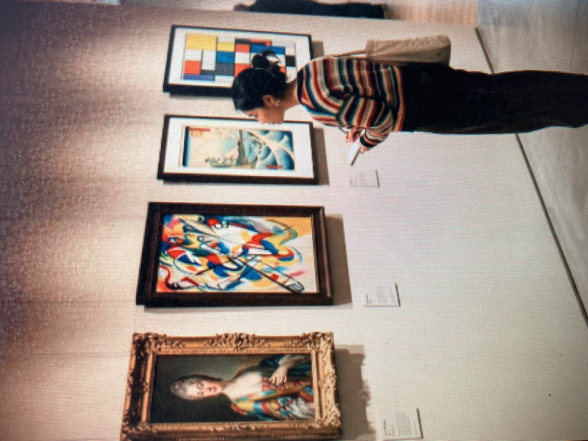In Sejahtera, Indonesia, a woman stands in a museum gallery, observing a collection of paintings. She is positioned to the right of the frame, facing left, and appears to be taking notes on a small pad. She wears a horizontally striped sweater in red, blue, yellow, and white, and dark trousers. A canvas tote bag hangs from her right shoulder.
The gallery wall is a neutral, textured color, and four framed artworks are displayed. On the far left, a portrait of a woman in a colorful, ornate dress is visible in an ornate gold frame. To its right is an abstract painting with bold, swirling lines of red, blue, yellow, and white. The third artwork is a landscape painting depicting mountains and water in shades of blue and white. The fourth painting, on the far right, is a geometric composition of colored squares and rectangles in red, blue, yellow, and white. Small white labels are affixed to the wall below each artwork. The lighting in the gallery is subdued, casting faint shadows on the wall. The floor appears to be polished concrete.