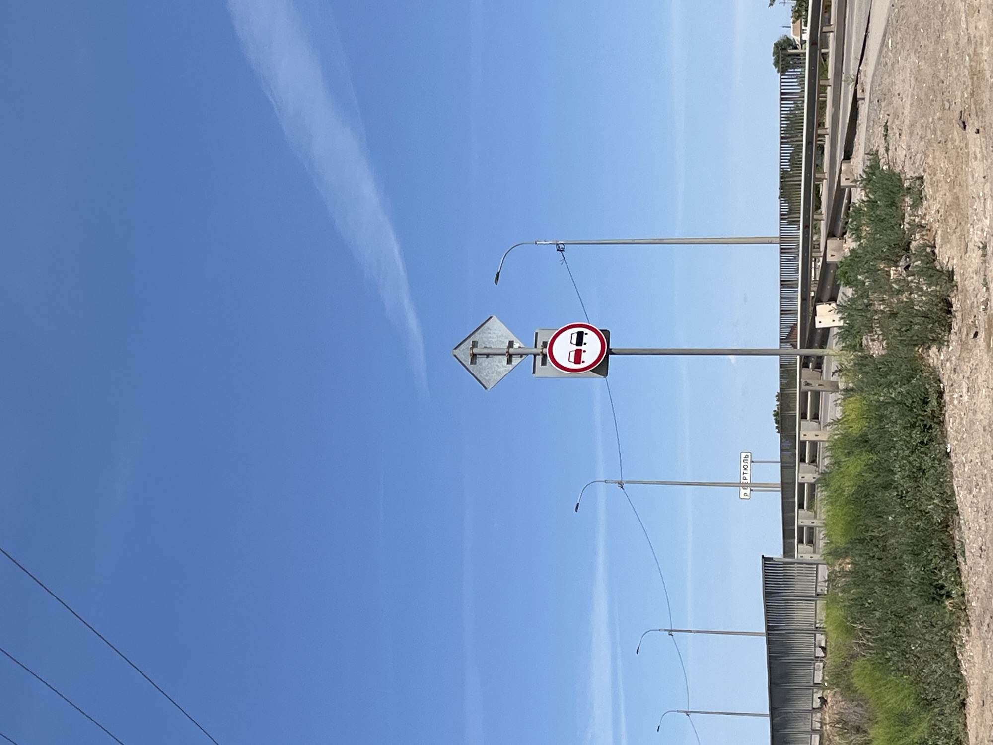 The image shows a road signpost on a clear day in Krasnyye Barrikady, Russia. The signpost has two signs: a diamond-shaped sign at the top and a circular sign below it. The circular sign is red and white and depicts two cars, indicating a prohibition of overtaking. The road ahead appears to be a bridge or overpass with a metal railing and streetlights. In the background, there is a white sign with Russian text, which appears to be a place name. The foreground is a dusty, unpaved area with sparse green vegetation. The sky is a vibrant blue with a faint cloud streak.