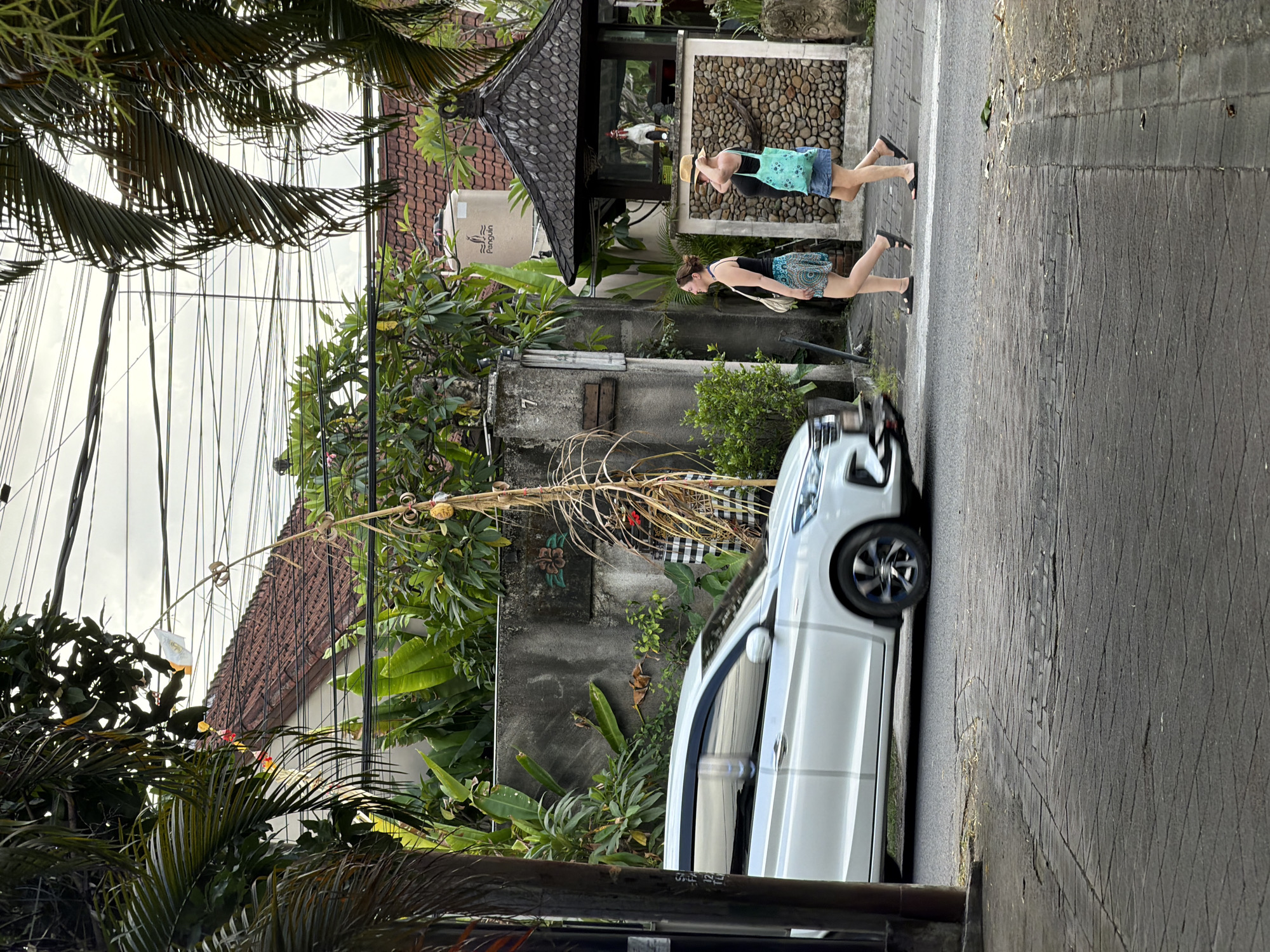In Kuta, Indonesia, a white vehicle is captured in motion on a street paved with interlocking bricks. Two women are walking on the sidewalk, one wearing a black tank top and a patterned skirt, the other a black tank top, a denim skirt, and a straw hat. A tall bamboo structure adorned with a white flag and yellow fruit stands near a stone wall covered in lush greenery. A gazebo with a thatched roof, built with stones and wood, is also visible in the background, and within it, a white rooster is standing. A water tank with the "Penguin" logo is positioned on the roof of a nearby building. Overhead, a complex network of electrical wires crisscrosses the sky.