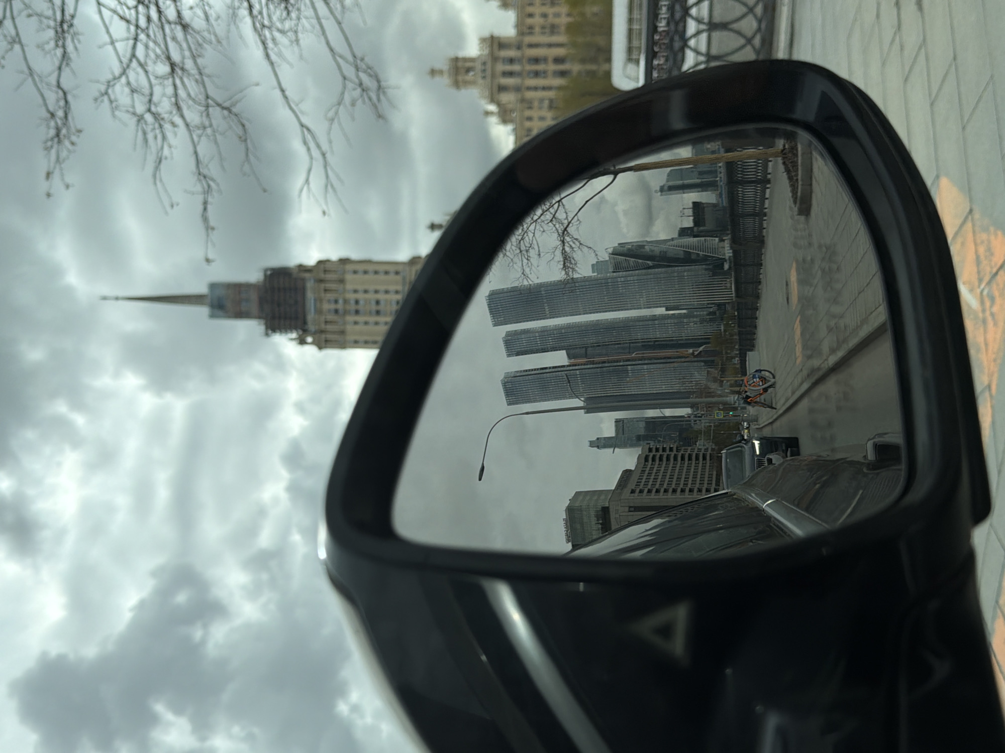 The image shows the reflection in a car's side mirror of the Moscow International Business Center (Moscow-City), Moscow, Russia. A classical Stalinist skyscraper can also be seen. Bare tree branches are visible against a cloudy sky. A cyclist is visible in the mirror's reflection. The immediate foreground is the car itself, including the side mirror. The road surface is partially visible.
