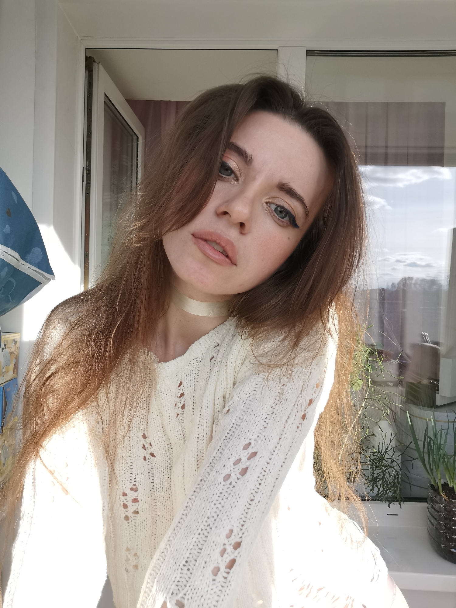A young woman with long brown hair is posing for a selfie in front of a window. She is wearing a white knit sweater and a light-colored choker. The background shows a window with visible plants on the windowsill and a partial view of what appears to be an urban landscape outside. The location is Unknown, Unknown.