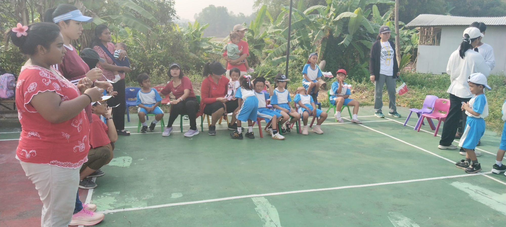 In Taban, Indonesia, a group of children and adults are gathered on a green outdoor court. The children, dressed in blue and white uniforms, are seated on colorful chairs, some looking expectant, others appearing tired. Several adults, presumably parents or guardians, stand behind them, some holding infants. One woman in a red shirt and light-colored pants stands in the foreground, holding what appears to be a spoon. The scene suggests a school event or a children's competition, with the adults acting as supervisors or participants. The background features lush greenery and trees, indicating a rural or semi-rural setting.