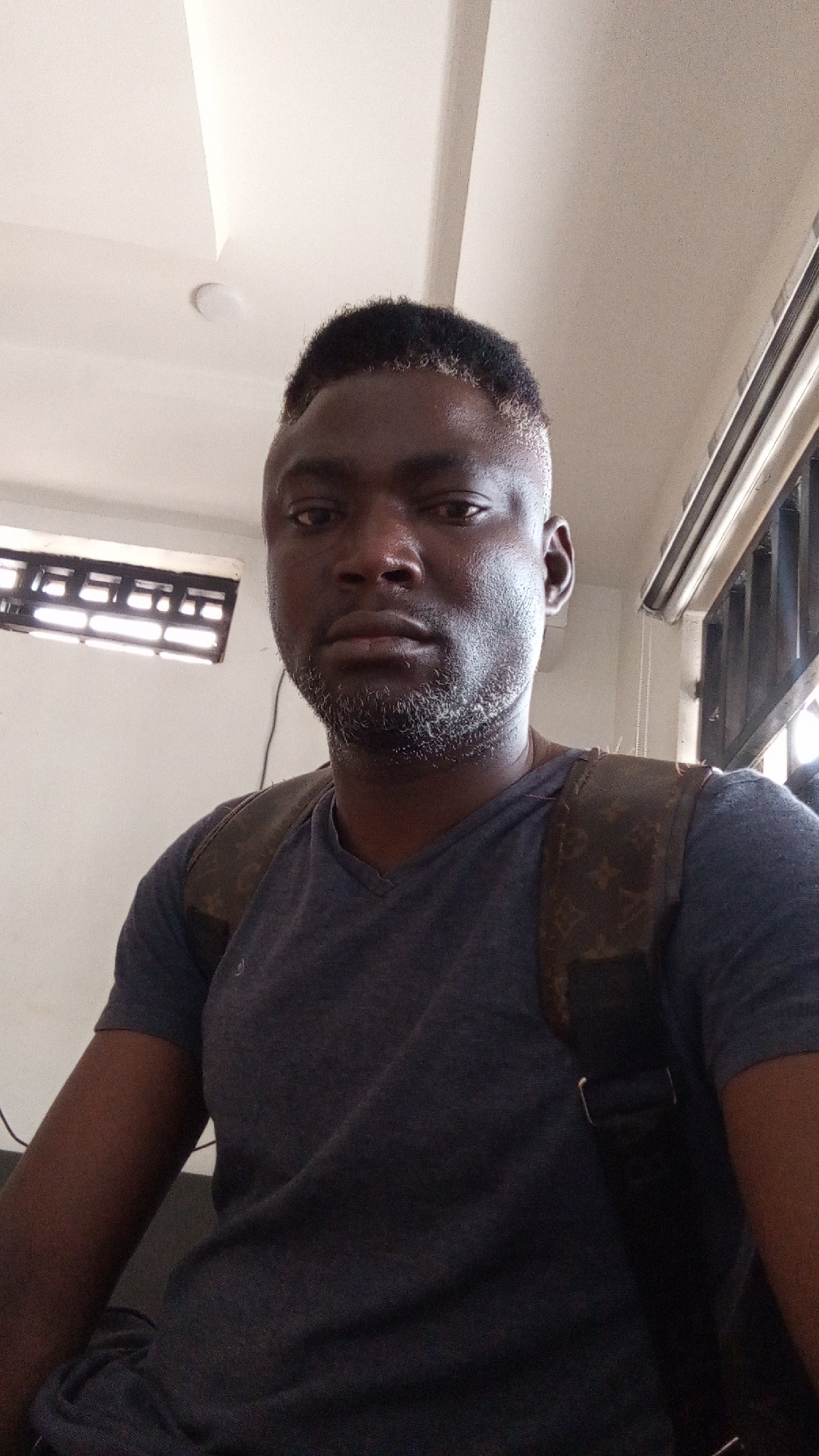 A close-up selfie captures a man with dark skin, short dark hair, and a neatly trimmed, salt-and-pepper beard. He is looking directly at the camera with a neutral expression. He wears a dark gray or blue V-neck t-shirt and a dark-colored backpack with a distinctive patterned design, possibly a luxury brand monogram, slung over his shoulders.
The setting appears to be indoors in Sardauna, Nigeria, with a simple, light-colored wall and ceiling visible in the background. A recessed ceiling light fixture is partially visible, and another lighter area suggests a window or another light source. To the left, a window with horizontal bars allows some light into the room. On the right, another window or opening with vertical dark bars is visible. The lighting is a mix of artificial and natural light. No specific activity beyond posing for the photograph is depicted. Wires or cables are faintly visible against the light wall on the left side of the frame.