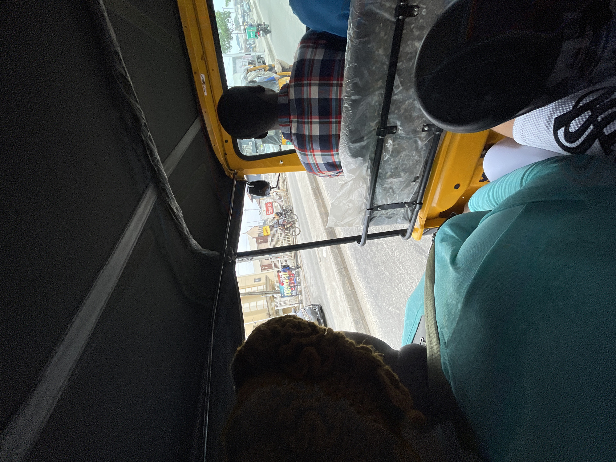 The image shows an interior view of a yellow auto-rickshaw, also known as a "Keke Napep," from the passenger seat. Two people are in the rickshaw, in addition to the driver, whose head is visible in the front. The rickshaw appears to be on a street in Ado, Nigeria, as buildings and street signs are visible through the front windshield. Specifically, the area is in Ado Ekiti.