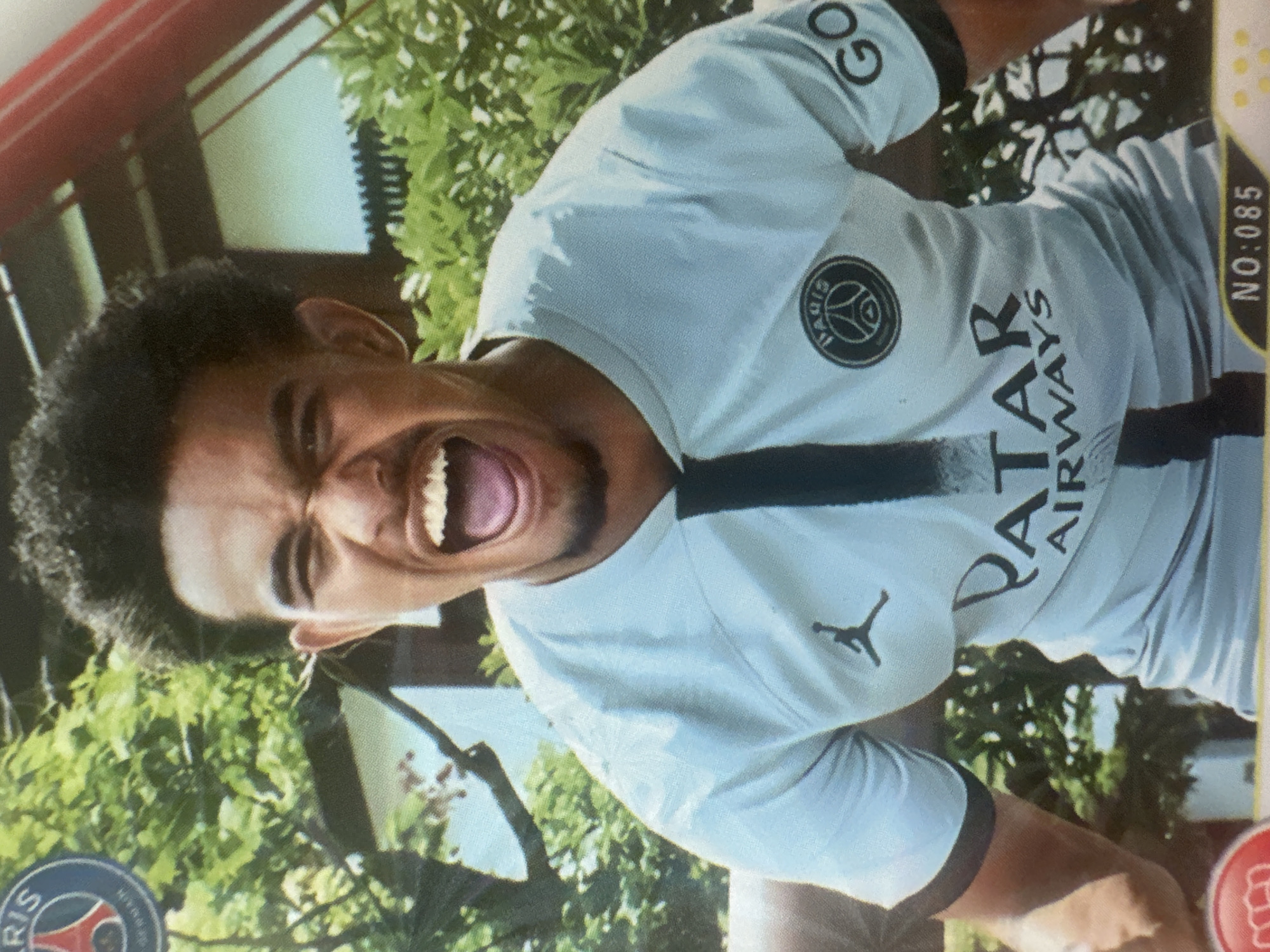The image shows a close-up of a young man wearing a light-colored soccer jersey with black accents. The jersey has several logos, including a circular crest with the word "PARIS" and an emblem, a Jumpman logo, and the word "GO" on the sleeve. Text on the jersey reads "QATAR AIRWAYS." The man has dark, curly hair and is shouting with his mouth open and tongue visible. He appears to be outdoors, with lush green foliage and what looks like a building in the background. A red border with a white icon and the text "NO: 085" are visible at the bottom. The image does not contain any information or context that would place it in the city of Minsk, Belarus.
