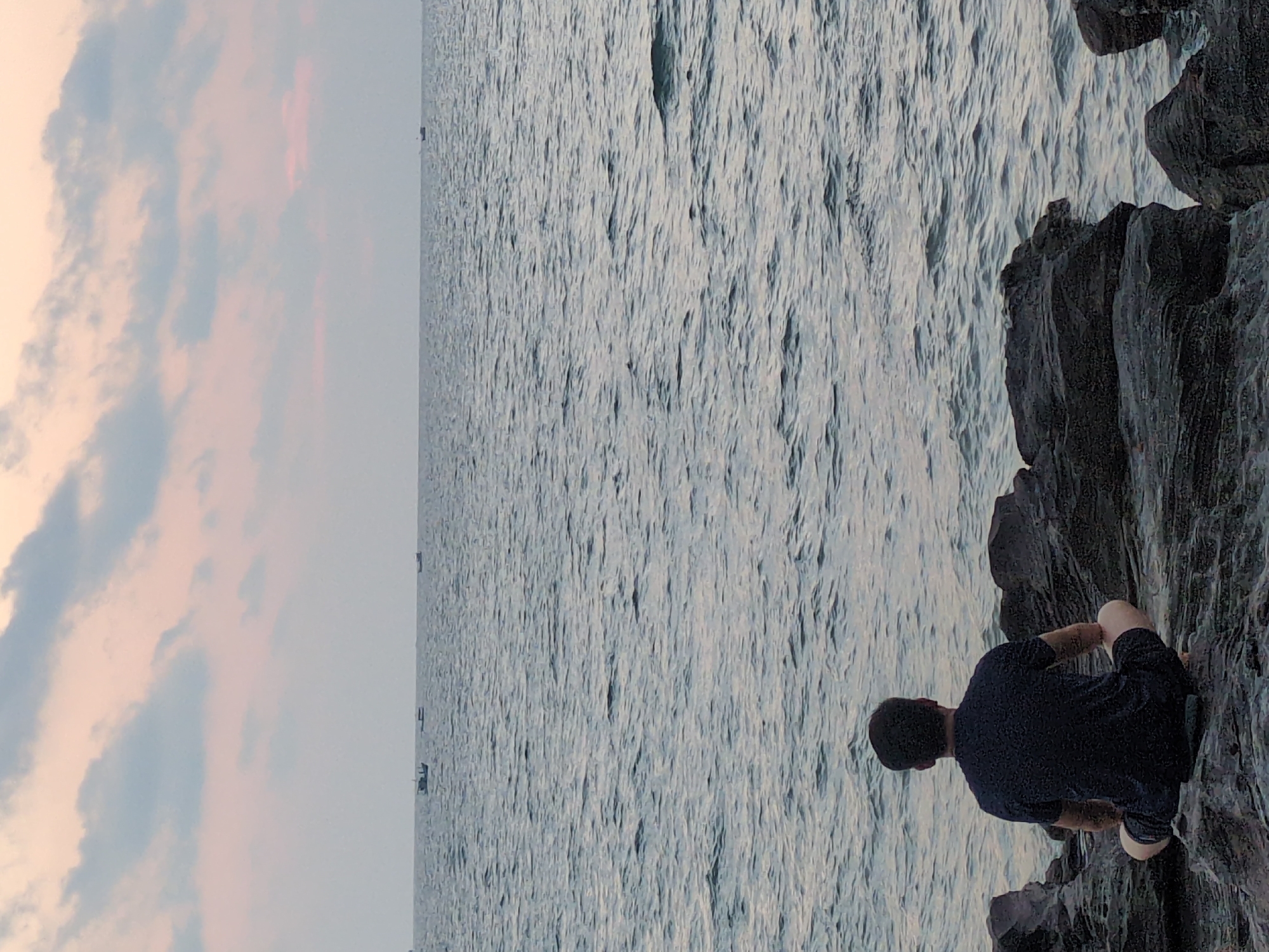A person is sitting on dark rocks at the edge of the ocean. The ocean stretches out to the horizon, with some small boats visible in the distance. The sky is partly cloudy, with a pinkish hue. The location appears to be a rocky coastline. The picture was most likely taken on Phú Quốc, Vietnam.