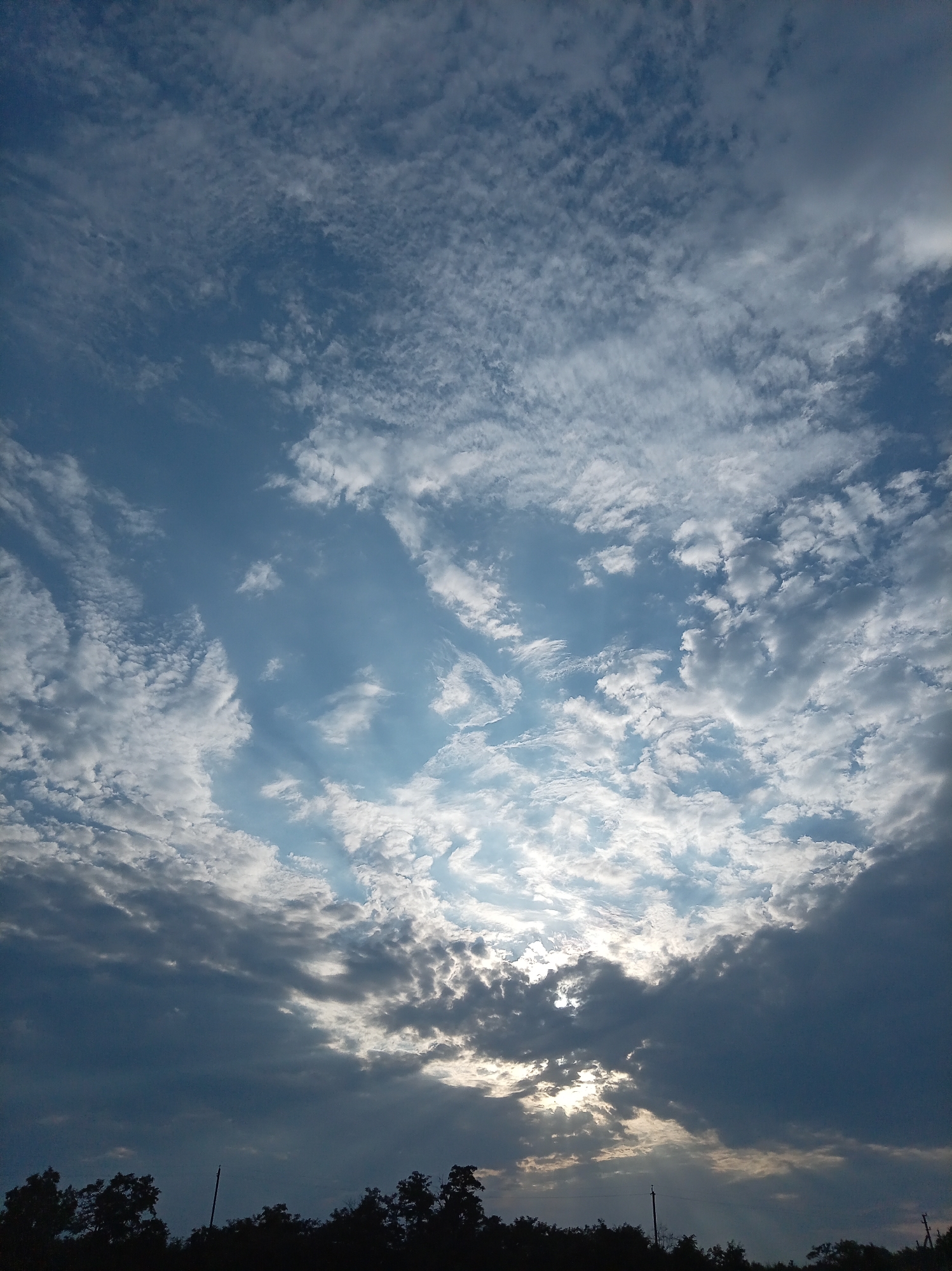 The image depicts a dramatic sky with a mix of dark and light clouds, with rays of sunlight piercing through gaps. The lower portion of the image shows a silhouetted tree line, suggesting an outdoor scene. The location in Гражданское, Russia, is not visually discernible from the provided image. There are no people or specific objects identifiable in the scene beyond the natural elements. The overall atmosphere is one of impending change, possibly due to the weather.