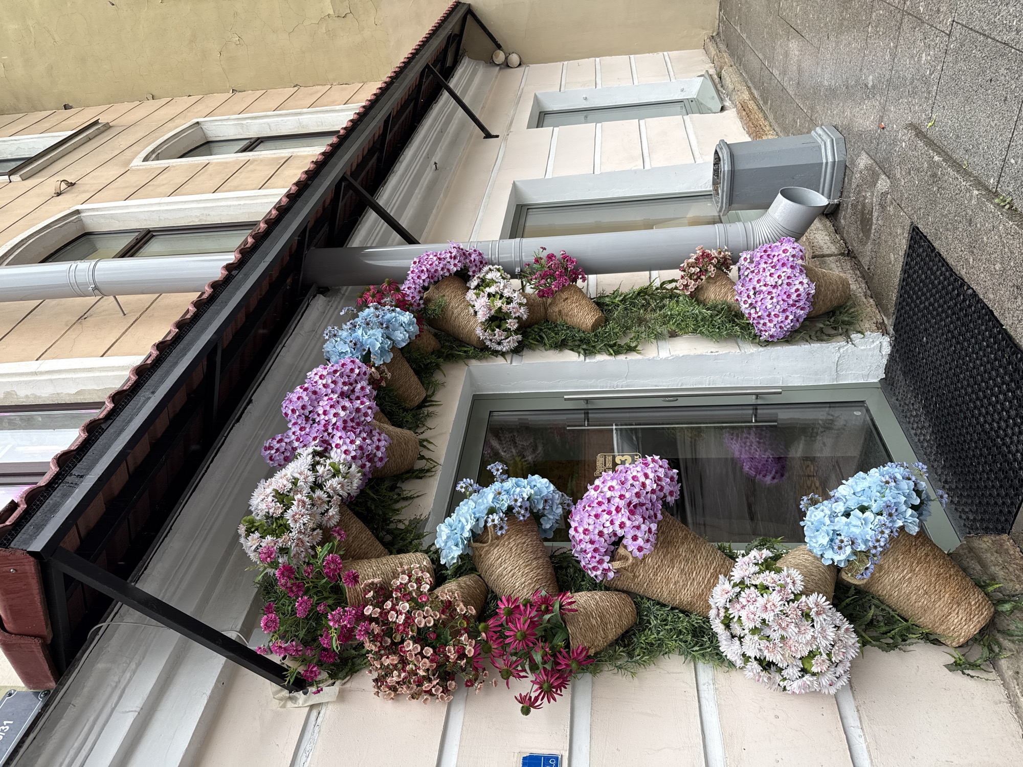 The image shows a building facade decorated with an array of flowers in woven containers. The building has beige walls with white trim around the windows. A gray drainage pipe runs horizontally across the front, and a roof overhang with dark trim and a dark support beam is visible. The flowers are various colors including purple, white, blue, and pink, and they are arranged in a garland-like manner around the window. The scene seems to be located in Saint Petersburg, Russia, as the building style and architecture align with the city's typical aesthetic. No people are visible in the image.