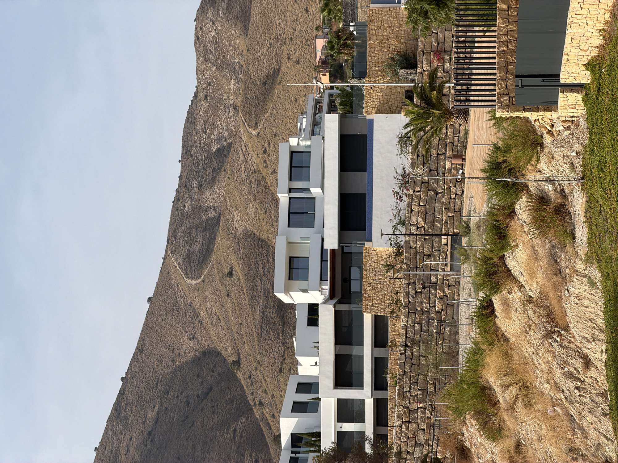 This image shows a modern, multi-story white building complex situated on a hillside in Finestrat, Spain. The buildings feature large, dark-paned windows and balconies with glass railings. The complex is built on a series of tiered levels, supported by stone walls. The foreground is a sloped, dry, rocky area with sparse, yellow-brown vegetation and some green bushes. A black metal fence is visible in the lower right corner. In the background, a dry, grassy hillside rises, with a faint, winding path visible on its slope. A few scattered trees are present on the hillside. The sky is a pale, clear blue. The scene appears to be a residential development in a Mediterranean climate.