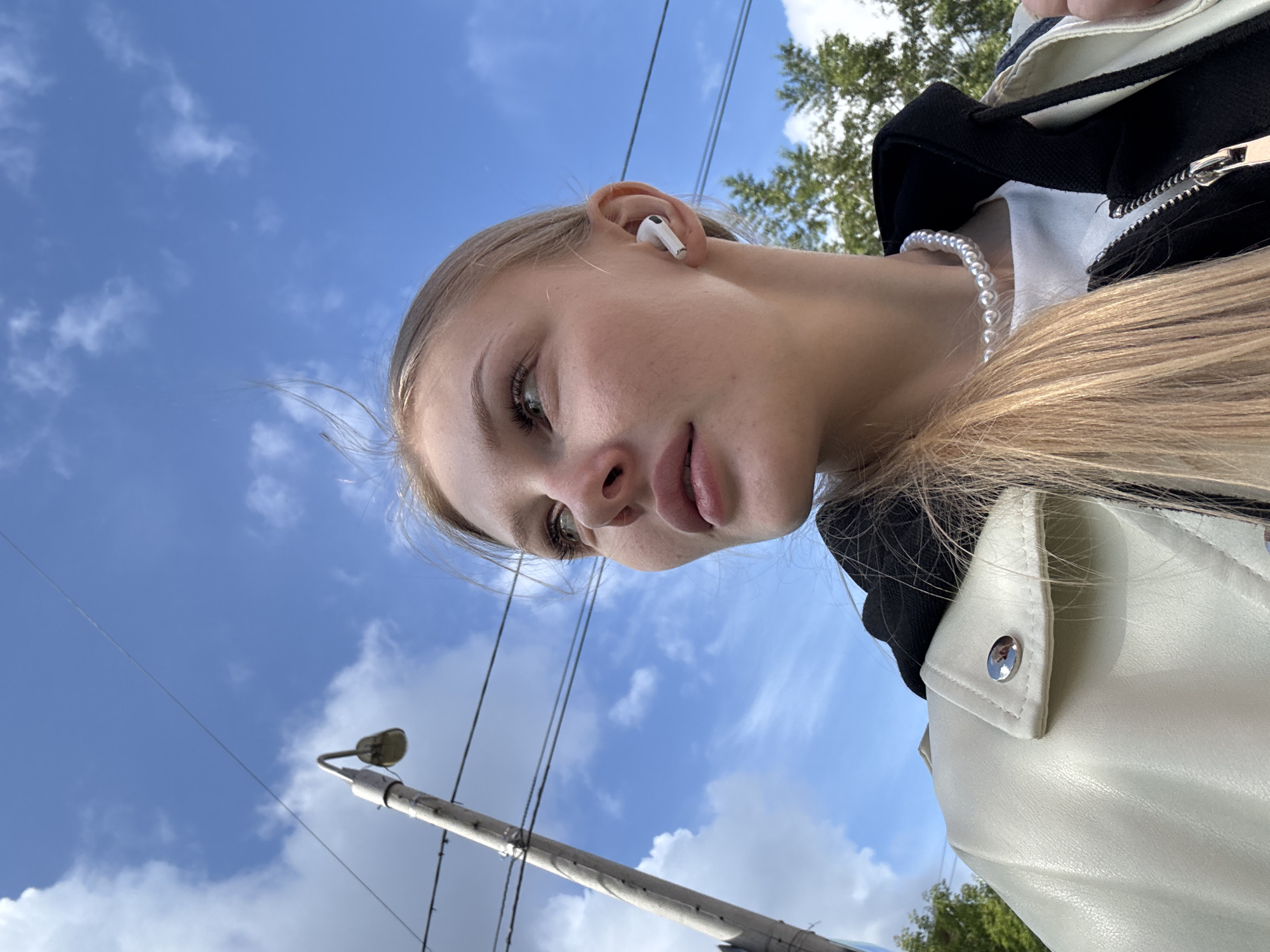 A young, fair-skinned woman with light blonde hair, appearing to be pulled back, is depicted in a low-angle shot, looking upwards and to her left. Her lips are slightly parted. A white wireless earbud (AirPod Pro) is visible in her right ear. She is wearing a cream-colored or off-white faux leather jacket over a black zippered hooded garment, which covers a white t-shirt. A pearl necklace is worn around her neck.
The background consists primarily of a bright blue sky with scattered white cumulus clouds. To the left of the frame, a grey utility pole with an attached street light fixture and multiple electrical wires runs vertically. Green foliage from trees is visible in the lower right and upper left background. The specific location in the city of Perm, Russia, cannot be determined from the visual information provided in the image.