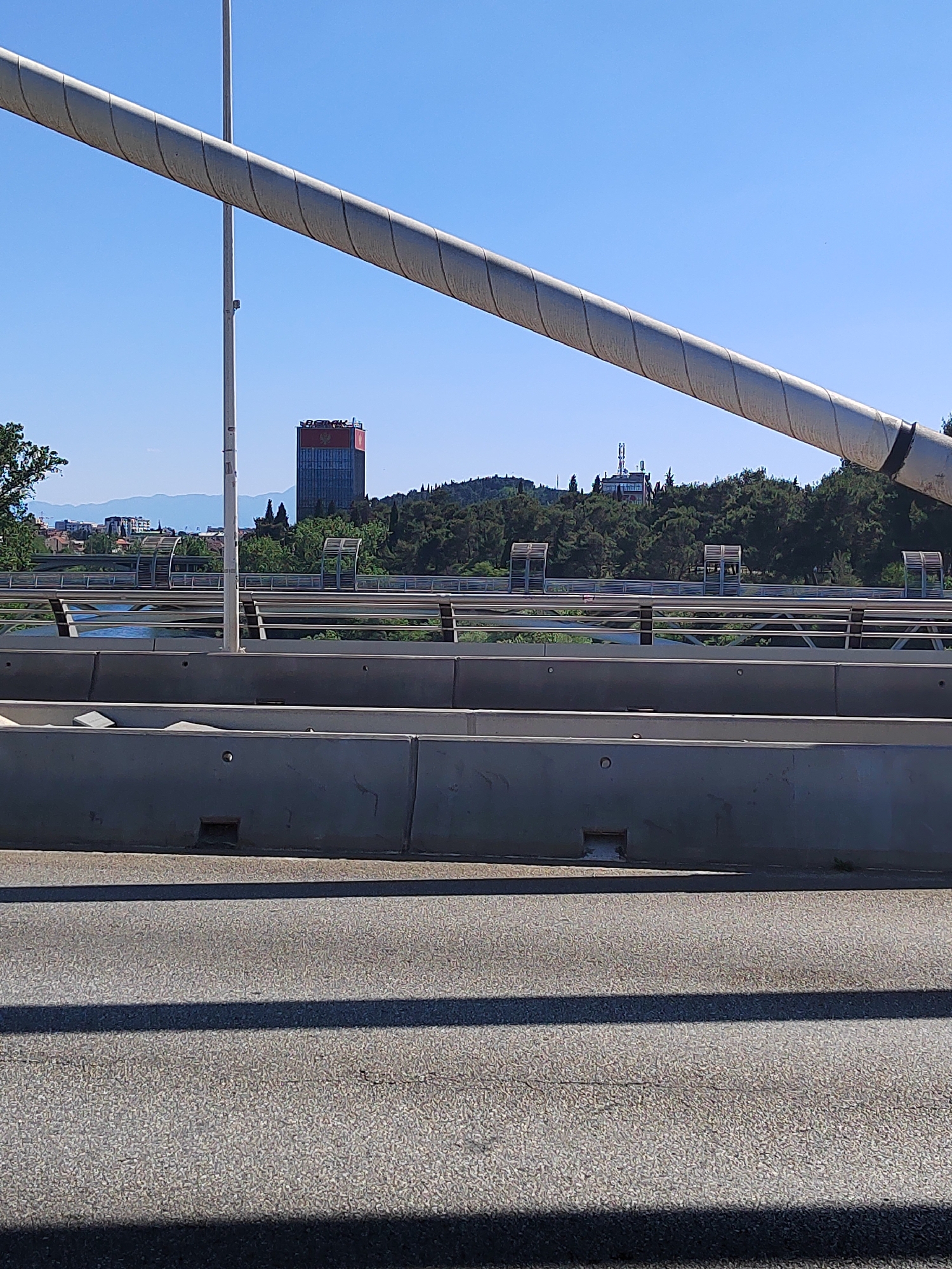 The image shows a section of a road or bridge in Podgorica, Montenegro. The foreground features an asphalt road surface with shadows. A concrete barrier is visible above the road, followed by a pedestrian bridge with metal railings. In the background, there's a view of the city, including the landmark building, The Capital Plaza, on a hill. Trees and other greenery are present in the middle ground. A large cylindrical structural component, possibly part of a bridge or support system, dominates the upper portion of the frame. The sky is clear and blue.