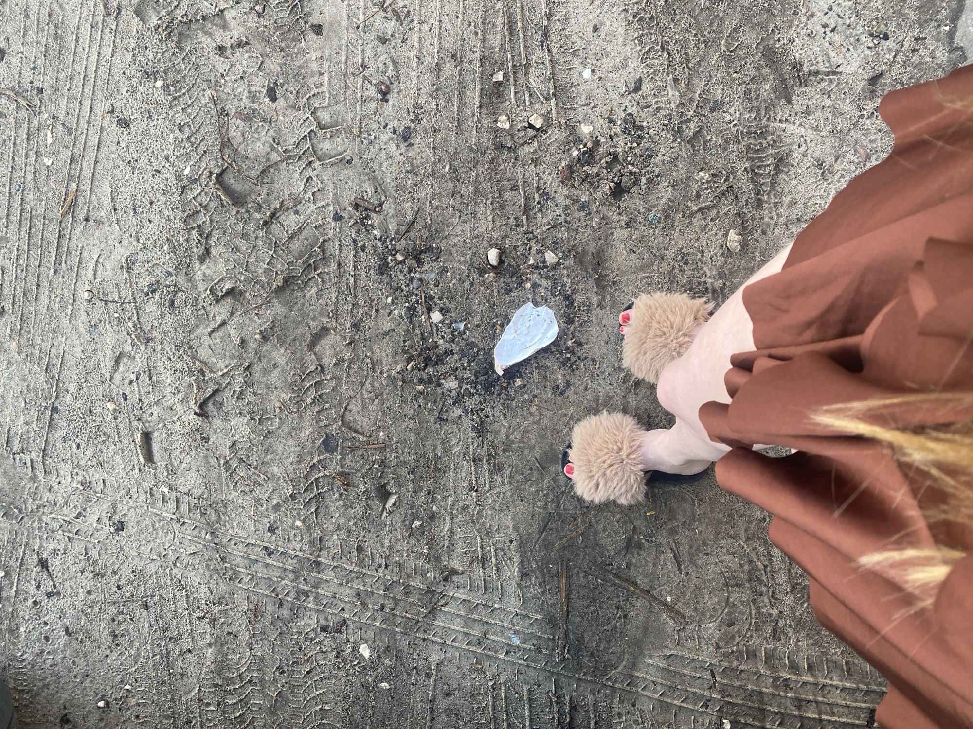 A person's legs and feet are visible from a top-down perspective, standing on a dirt ground with tire tracks imprinted. The person is wearing fuzzy brown slippers with red nail polish on their toes, and a brown dress. In front of the person, there is a piece of white plastic debris on the ground. The environment appears to be an outdoor area, possibly a dirt road or parking lot, with small stones and twigs scattered on the surface. The tire tracks suggest vehicular activity in the location. No specific landmarks of Медведево, Russia are identifiable.