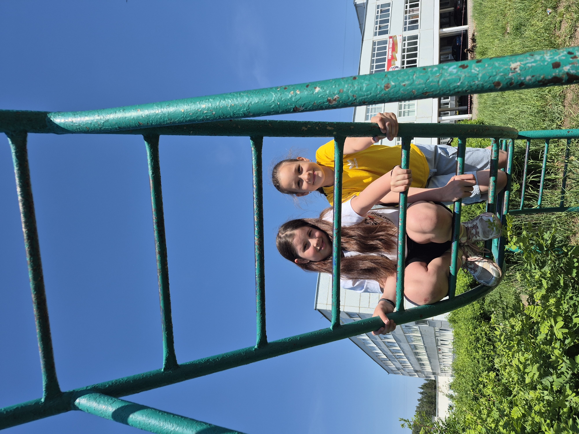 Two girls are on a green metal playground ladder. The girl in the foreground has long brown hair, is wearing a white shirt and dark shorts, and is squatting on the ladder. The other girl is wearing a yellow shirt and is behind her. Behind the ladder, there is a multi-story white building with windows. The sky is blue. Green foliage is visible on the ground near the building.