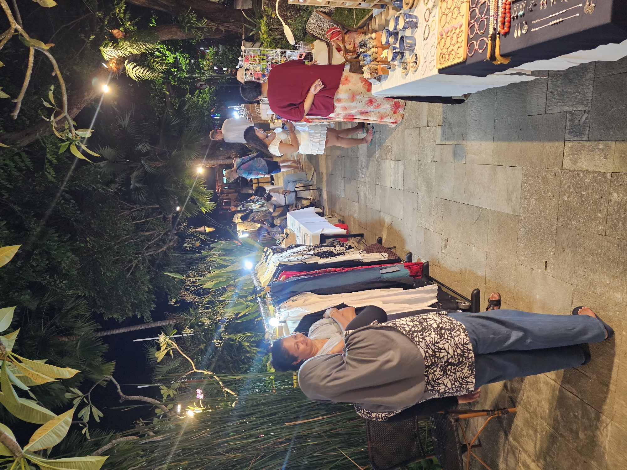 The image shows a nighttime scene in Denpasar, Indonesia, likely at a street market. The environment features a stone walkway lined with stalls displaying items like clothing, jewelry, and ceramics. The area is illuminated by overhead lights interspersed among trees and foliage.
Several people are present, including vendors attending to their stalls and customers browsing the goods. A woman is seated on a chair near a rack of clothes, resting. Others are actively engaged in looking at the merchandise, while a child in a white dress stands with an adult examining a display of ceramics and jewelry. Interactions consist of shopping and vendor-customer activity.
The objects of interest are the merchandise stalls, clothing racks, jewelry displays, and ceramic items. The environment is a blend of urban street market and garden-like setting with trees and plants.