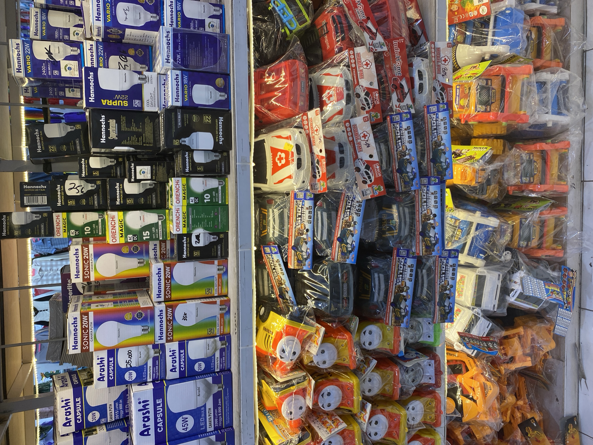 The image displays a retail environment, likely a store, exhibiting two distinct shelves stocked with merchandise. The upper shelf is predominantly filled with boxes of light bulbs, featuring brands like "Arashi," "Hannochs," and "Orenchi," with wattage and type specifications clearly visible. The lower shelf showcases a variety of toys, including miniature vehicles such as police cars, ambulances, construction trucks, and toy trains, all individually packaged in clear plastic. The overall setting suggests a commercial display designed for customer selection. The background is blurred, but hints at clothing items on hangers, possibly indicating a larger general store or department store. The environment is well-lit, facilitating a clear view of the products.