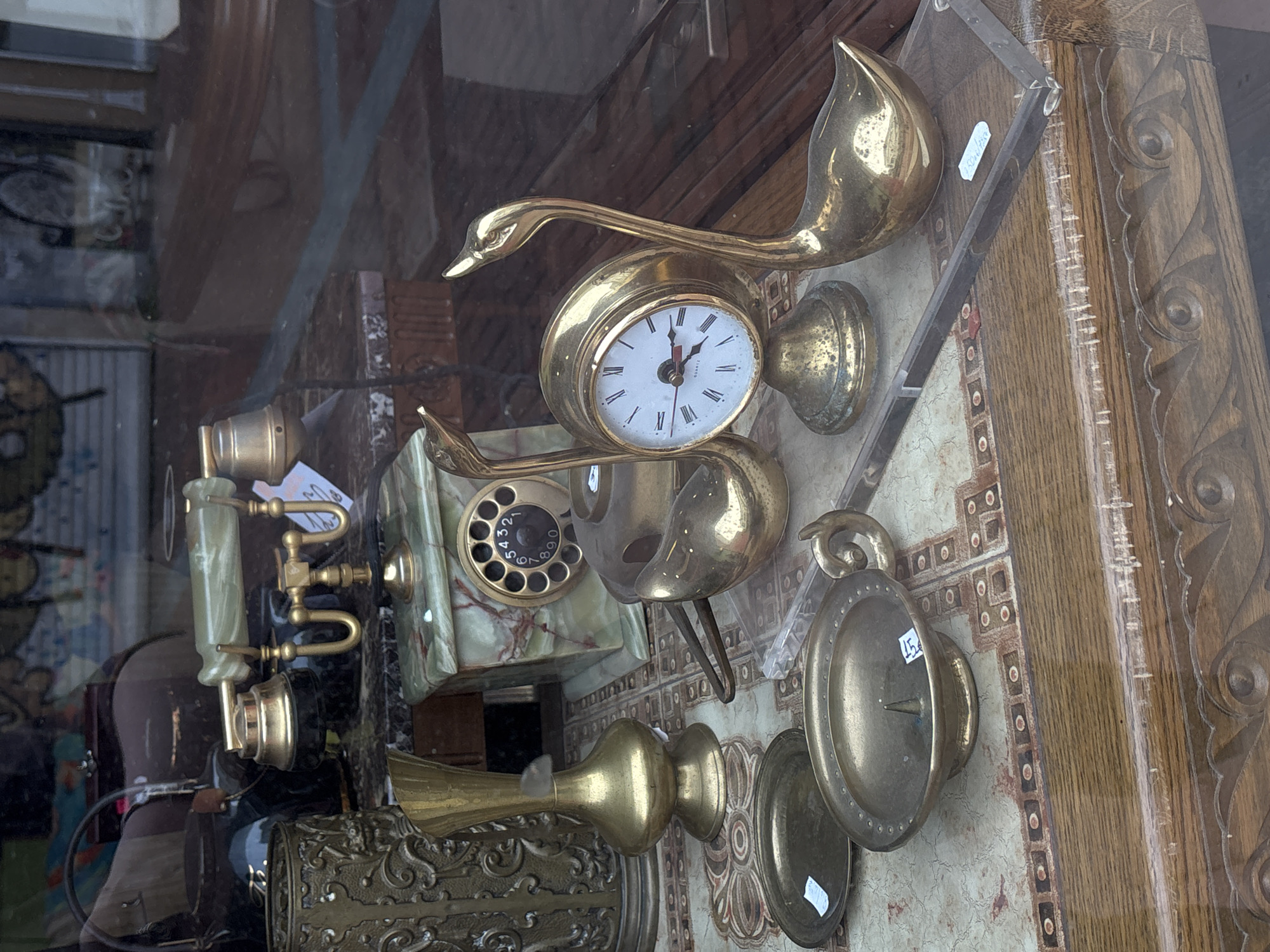 The image shows a display of antique objects in a shop window in Varna, Bulgaria. A gold-colored swan clock sits atop a patterned surface, with a green and black rotary telephone to its left. A brass candle holder is positioned near the right edge. The background features a reflection of the street outside, blurring the interior and exterior environments.