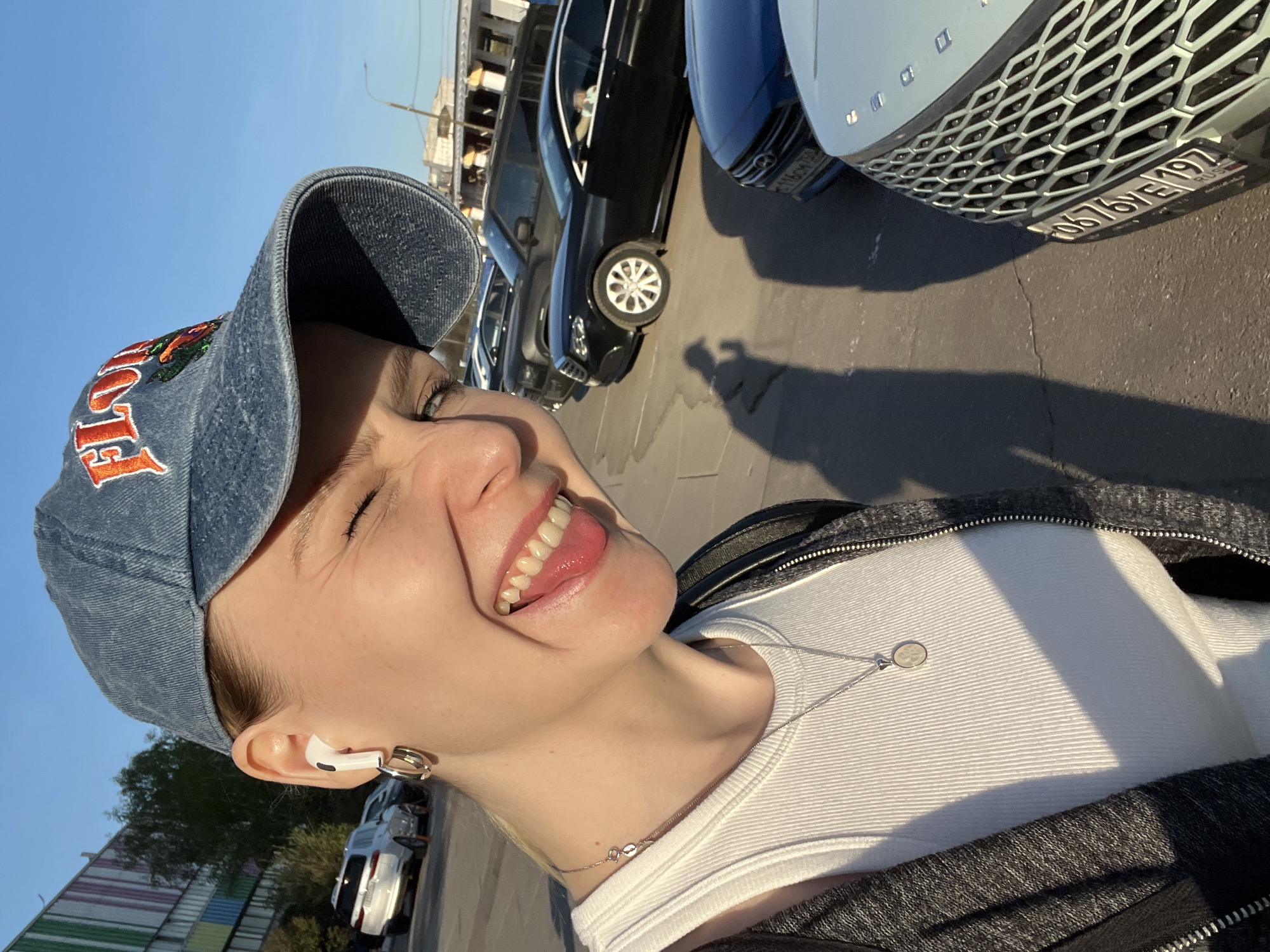 A young woman is depicted in a close-up selfie. She is wearing a denim baseball cap, a white tank top under a dark jacket, and a silver necklace with a circular pendant. She is also wearing an earpiece. The woman is smiling widely, her eyes are closed, and her teeth and tongue are visible.
Behind her, a row of parked cars can be seen. The license plate on the vehicle closest to the camera reads "O676YE 197 RUS".
The environment seems to be an urban setting with buildings visible in the background and along the periphery of the image. Considering the license plate ending in "197 RUS" is associated with the city of Moscow and the surrounding Moscow Oblast, the scene is likely set in or around Khimki, Russia. The lighting suggests it is daytime.