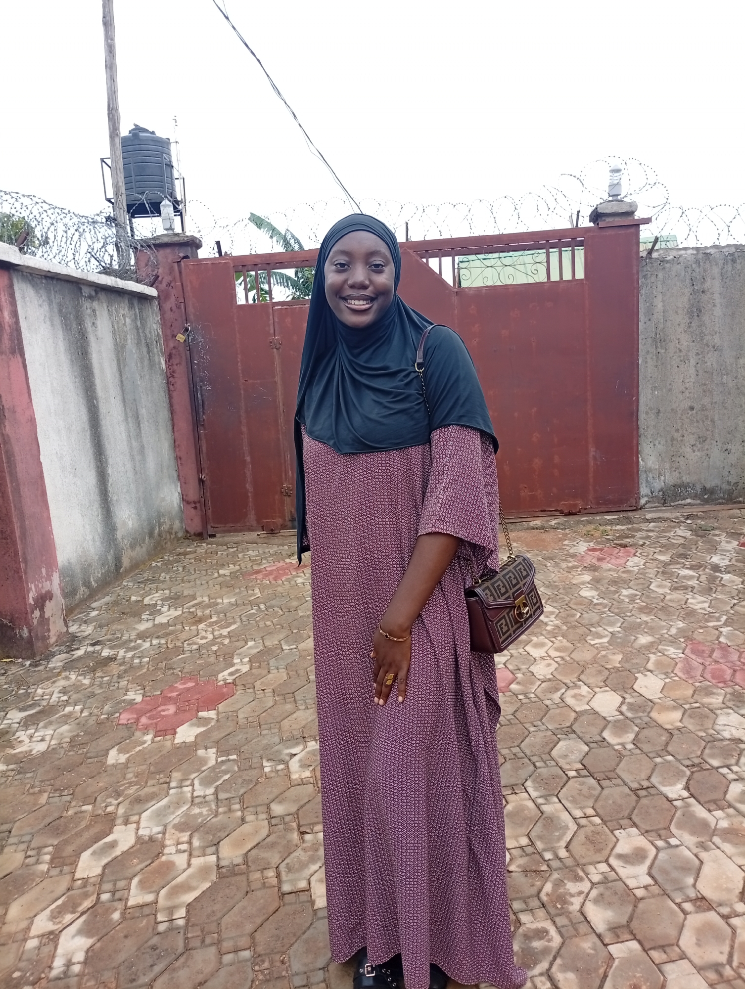 A woman stands outdoors in Rido, Nigeria. She wears a black hijab and a long, patterned dress. A brown handbag hangs from her shoulder. Behind her is a red metal gate topped with barbed wire, flanked by concrete walls. A water tank is visible on a pole next to the gate. The ground is paved with geometric stones.