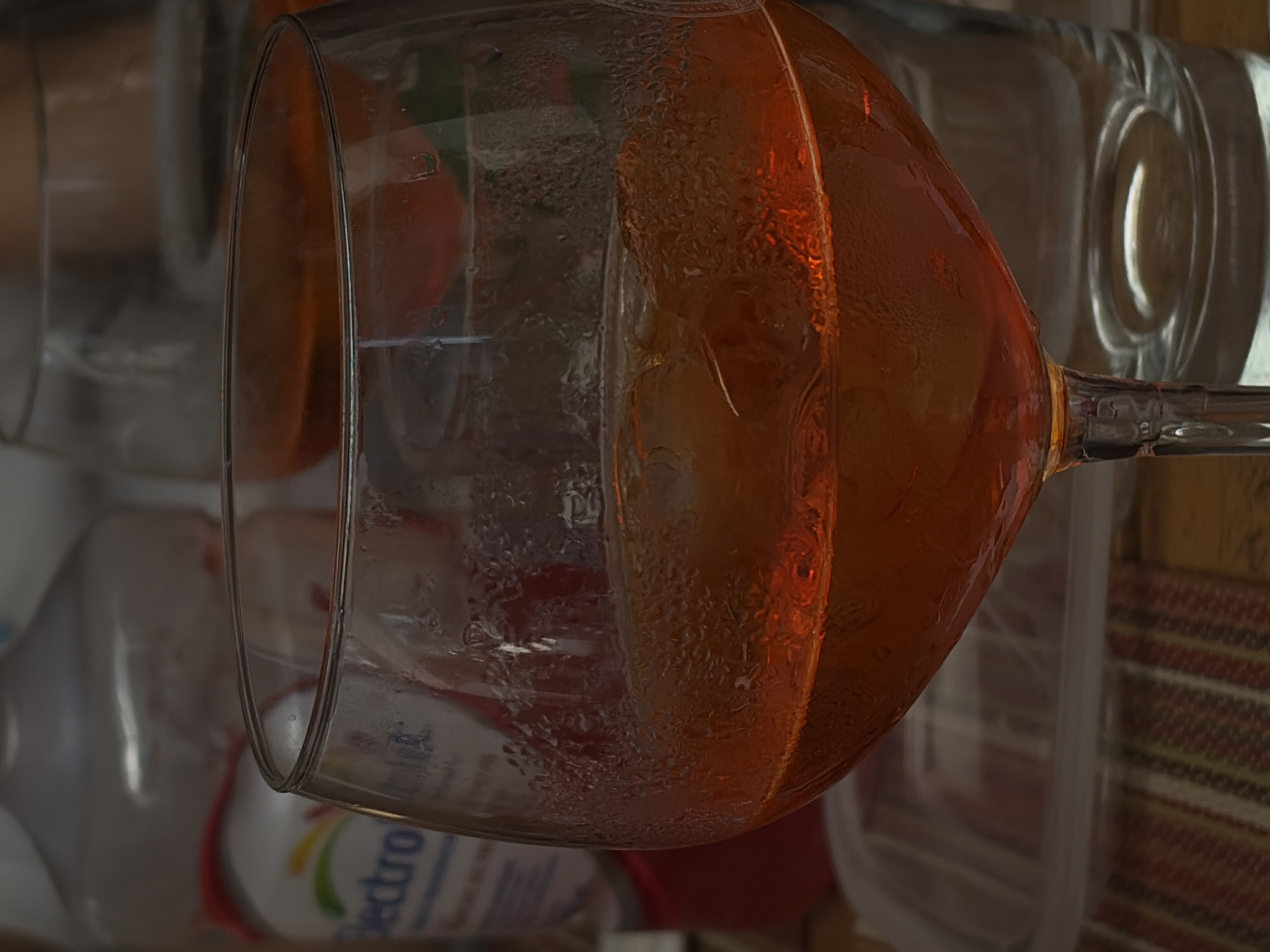 A close-up, tilted shot focuses on a partially-filled wine glass. The glass contains a light brown liquid with condensation on the outer surface. Several other glasses are visible in the background, along with a bottle labeled "ectro" and a red object. The overall setting seems indoors, possibly a kitchen or storage area. The image does not inherently suggest a specific location in San Bernardino Tlaxcalancingo, Mexico.