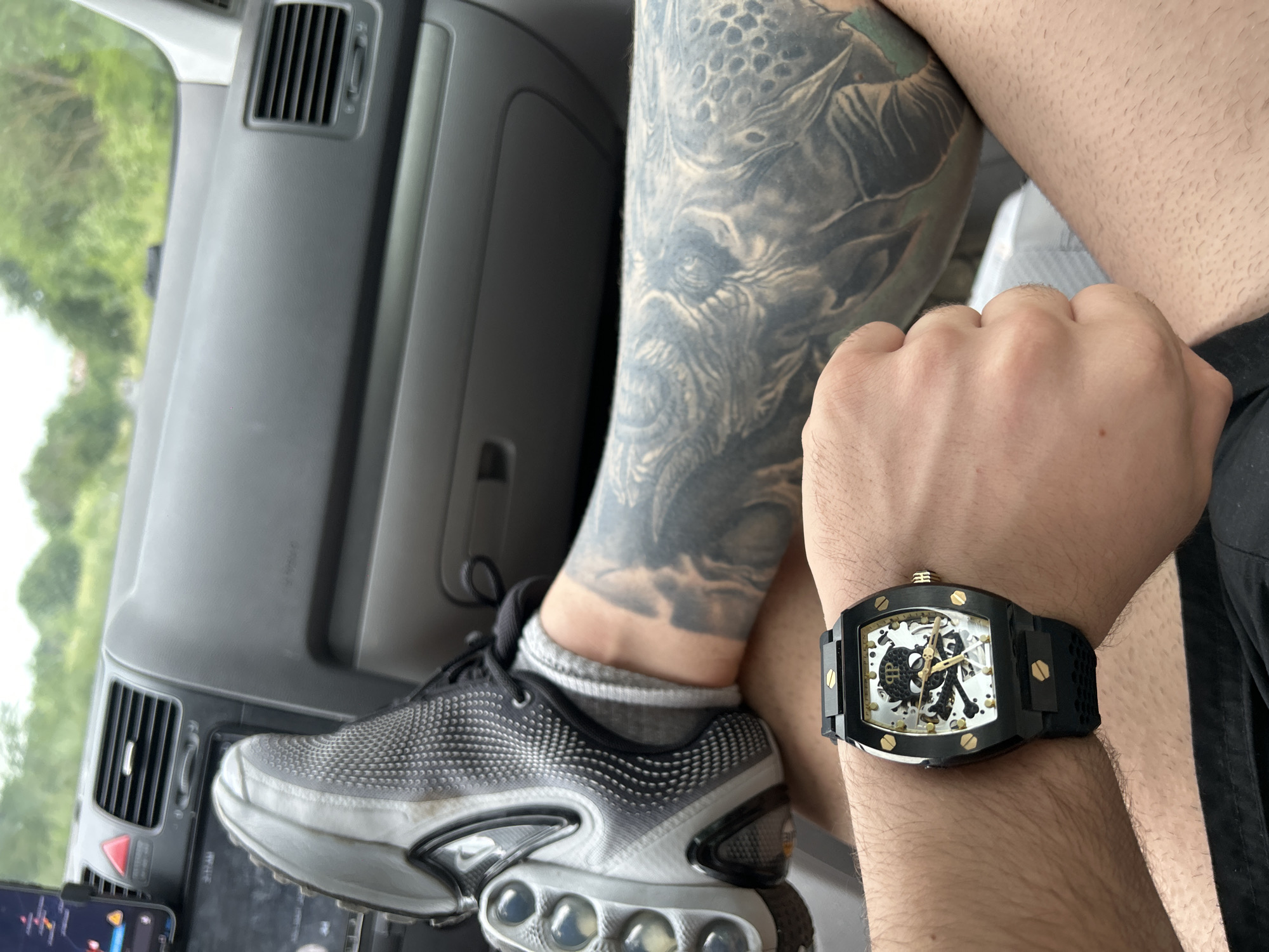 A first-person perspective image shows a person seated inside a vehicle, likely a car, with a tattoo on their left leg and a watch on their left wrist.
The leg, heavily tattooed with a grayscale design depicting a monstrous figure, is positioned in the foreground. A black and gray Nike Air Max Plus sneaker is visible on the foot. The shoe rests on the dashboard, partially obscuring the car's interior features, including an air vent and a touchscreen interface.
The left wrist displays a black and gold skeleton-themed watch with a skull and crossbones motif on the face. The watch band appears to be made of a textured black material.
The vehicle's interior consists of gray plastic components. Outside the car's windshield, green foliage is visible, suggesting a natural environment.
There is no evidence that indicates the location to be Medovo, Bulgaria.