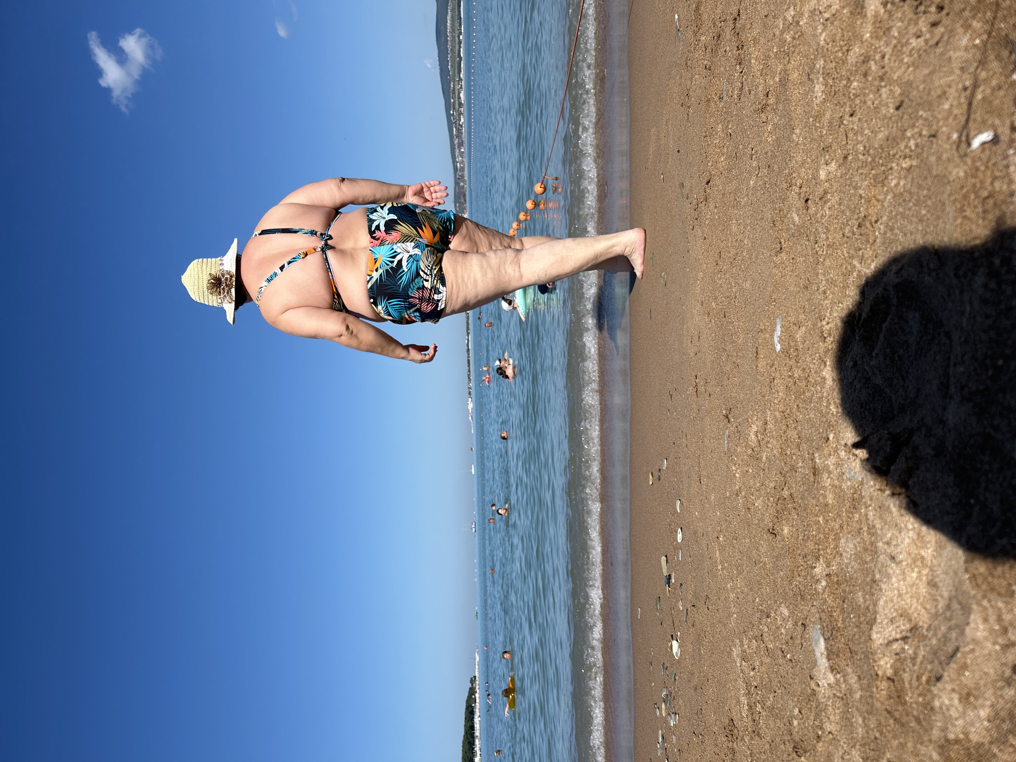A woman wearing a floral-patterned bikini and a straw hat stands on a sandy beach, facing away from the camera. She is looking out at the sea where several people are swimming and some boats are visible in the distance. The water is calm with small waves lapping at the shore. The sky is clear and blue, with a few scattered clouds. The scene appears to be a sunny day at the beach in Gelendzhik, Russia.