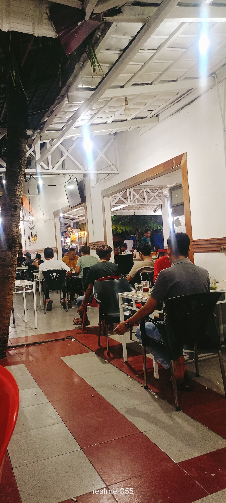 The image depicts an indoor/outdoor cafe setting at night. Several patrons are seated at tables, some facing away from the camera. The cafe appears to have a covered outdoor seating area with a white, slatted roof and exposed wooden beams. There is a tall palm tree trunk on the left side of the image. The floor is tiled with a red and white checkered pattern. A television screen is visible mounted on the wall. The cafe is illuminated by artificial lighting, including exposed bulbs and bright, blurry light sources that suggest flash or camera glare. Some patrons are engaged in conversation or eating. The overall atmosphere suggests a casual dining or gathering place.
