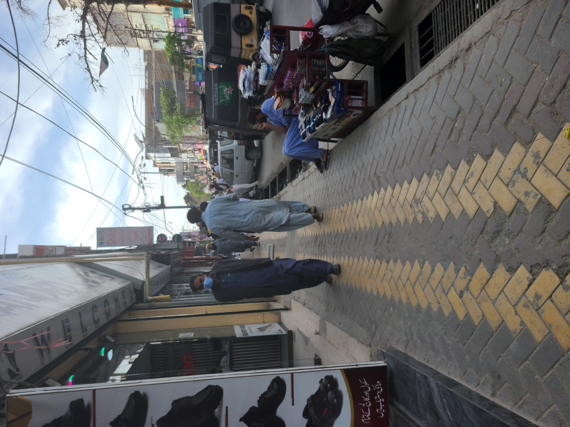 The image depicts a street scene in Quetta City Tehsil, Pakistan. The street is lined with shops, and people are walking along the sidewalk. There are vendors selling goods on the side of the street. The sidewalk is made of patterned concrete bricks. Utility lines are visible overhead. The sky is partly cloudy.