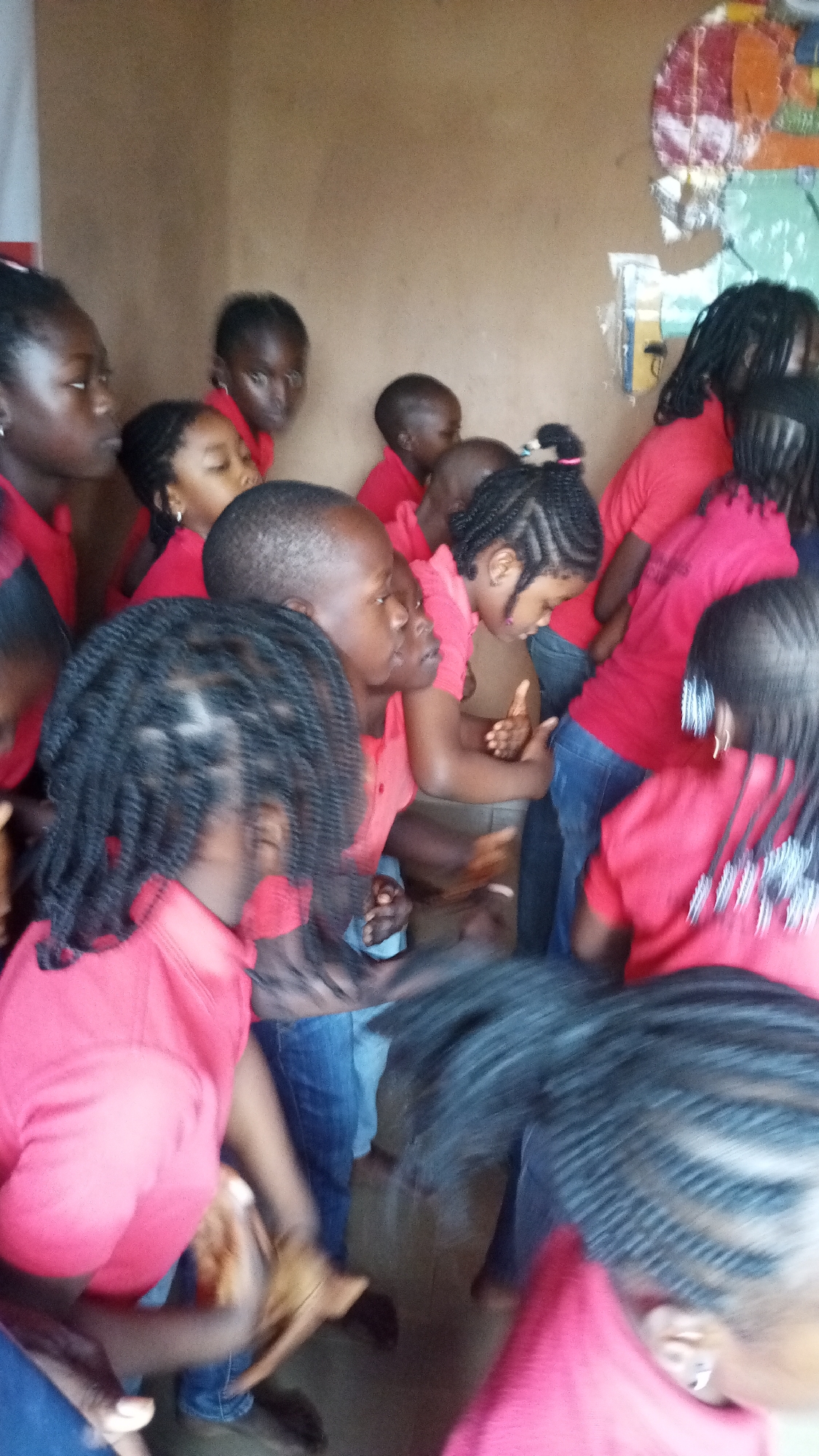 In an indoor scene with earthen-toned walls, a group of children, all appearing to be under the age of ten, are clustered together, mostly wearing red tops and blue jeans. The children are predominantly of African descent, with varying hairstyles, including braided and dreadlocks. Their faces are mostly obscured or turned away from the camera, and their movements suggest activity, possibly dancing or playing, as evidenced by blurred limbs and gestures. Some children are looking towards the right side of the frame, while others are facing inwards. In the background, a piece of colorful artwork is partially visible on the wall, and some areas of the wall appear to be decorated with torn paper or fabric. The lighting is somewhat dim, casting soft shadows. There is no clear indication of the specific city or location within the city.
