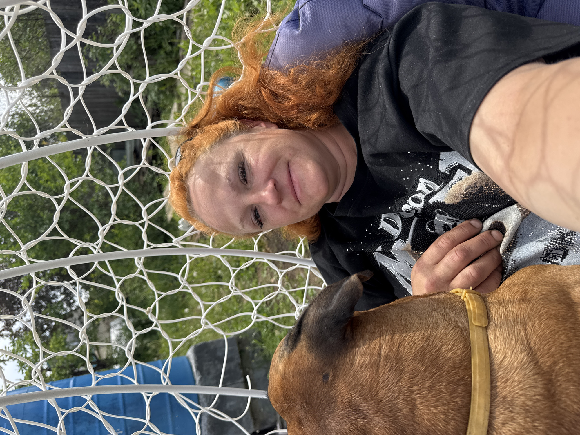 This outdoor selfie captures a woman and her dog in a relaxed moment, viewed through a white, decorative mesh structure, likely part of an outdoor chair or swing. The image is oriented on its side.
The woman, with vibrant red hair, is positioned on her left side, looking directly at the camera with a gentle, contented smile. She is wearing a black, short-sleeved t-shirt adorned with a sparkly design that appears to include the partial word "DOG" and an eye, hinting at a dog's face. Her right hand is visible, resting near her pet. Behind her head is a purple object, possibly a cushion or part of the chair.
Beside her is a brown, short-haired dog, seen from the back of its head and neck, wearing a light yellow/tan collar. Its ear is visible near the woman.
The background reveals an outdoor setting, likely a garden or backyard in Chaikovsky, Russia, with lush green foliage (trees and grass) under bright daylight. A blue object, possibly a barrel or a pool, is also partially visible. The scene conveys a sense of calm and affection between the woman and her dog.