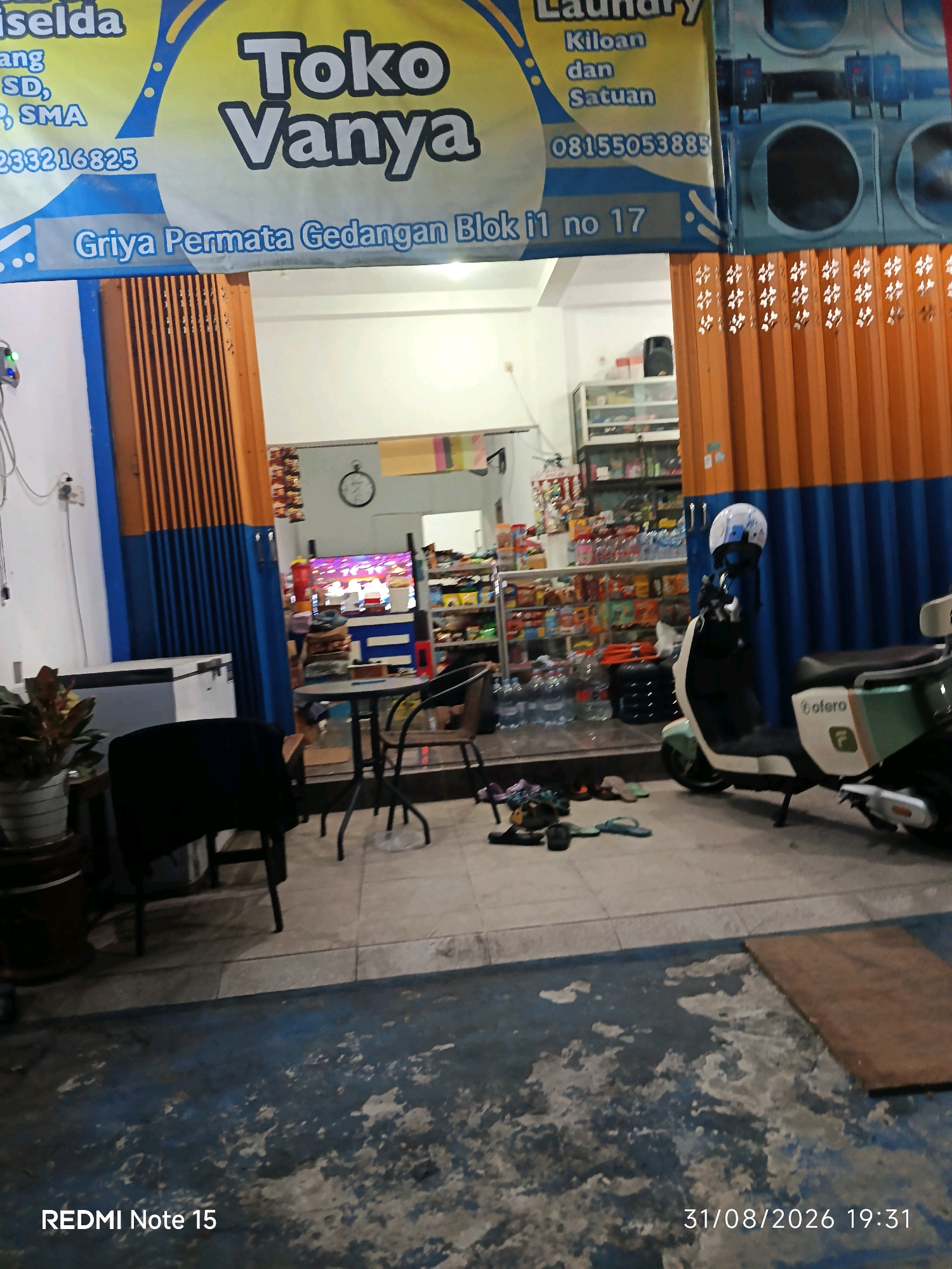The image shows the exterior of a small convenience store and laundry service named "Toko Vanya" located at Griya Permata Gedangan Blok i1 no 17 in Keboansikep, Indonesia. The storefront is covered by an orange and blue corrugated metal folding gate. The shop appears to be open, with products visible on shelves inside. A black chair and a round table are set up outside the entrance, suggesting a waiting or seating area. A white and green electric scooter is parked to the right of the entrance, with a white helmet on its handlebars. Various items are scattered on the ground in front of the shop, including footwear. The overall scene is that of a typical neighborhood business.