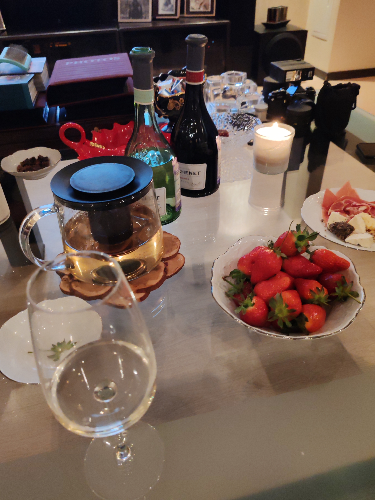 The image depicts a tabletop still life. A glass of clear liquid, possibly wine, stands in the foreground, slightly out of focus. Next to it is a small white plate. A glass teapot filled with tea sits on a wooden coaster. Adjacent is a small white bowl containing dark-colored objects, possibly dates or dried fruit. Two wine bottles, one green and one dark red, are positioned behind the teapot. In front of the bottles is a red teapot-shaped object. Further to the right, a lit white candle stands in a glass holder. A bowl of strawberries and a plate with cheese and meat are also visible on the table. In the background, there's what appears to be a bookshelf with books and photo frames. There is no evidence to confirm this location as Saint Petersburg, Russia. The scene seems to depict a private or domestic setting with food and drinks arranged on a reflective surface.
