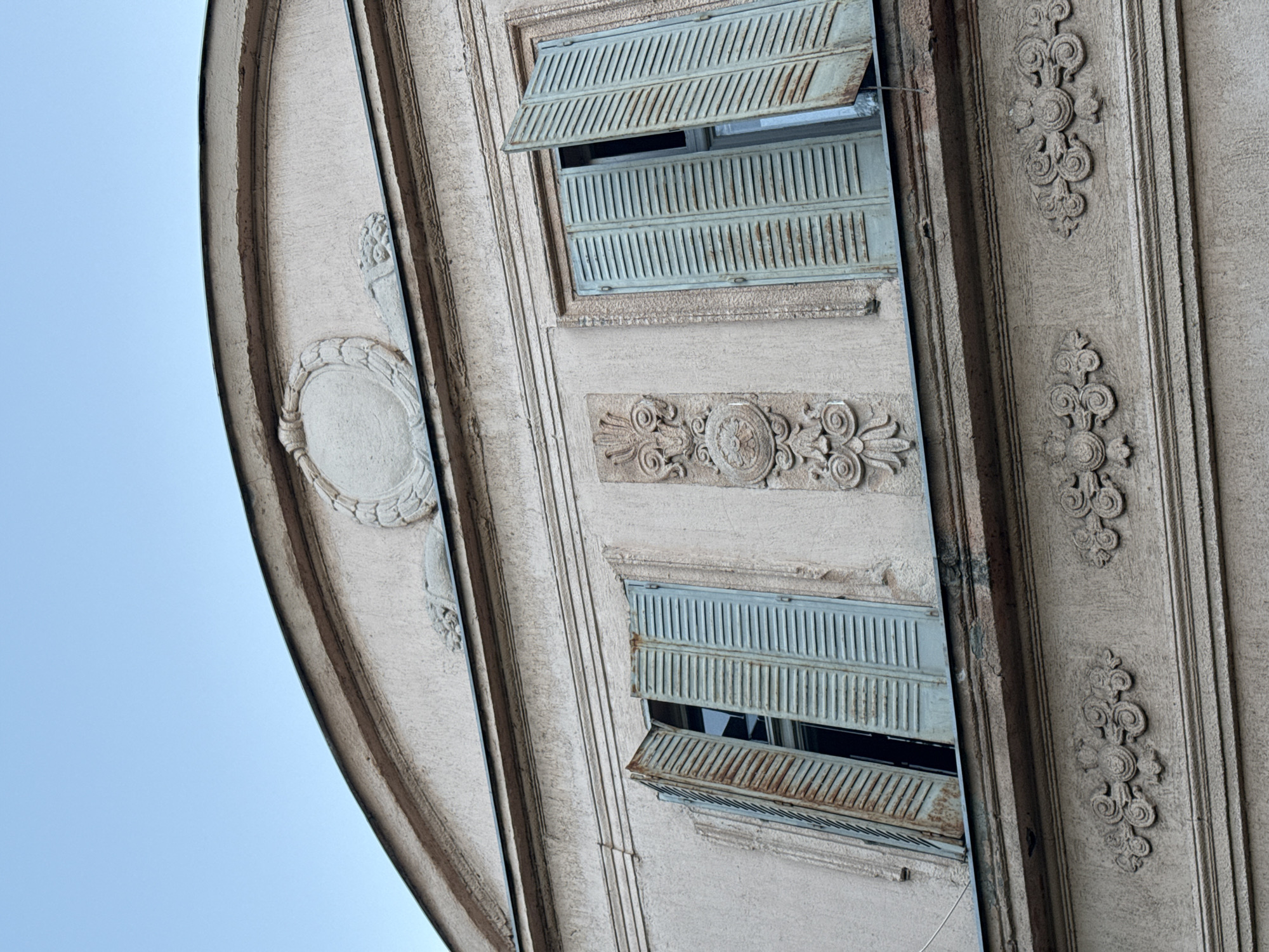 A low-angle shot captures a portion of a building in Vracar Urban Municipality, Serbia. The building features light-colored facade with ornate embellishments, including raised oval designs and floral-inspired patterns. Two windows with slatted, light-green shutters are partially open, revealing a dark interior. The shutters show signs of aging with some rust evident on the bottom window's lower shutter. The sky is visible in the left portion of the image.