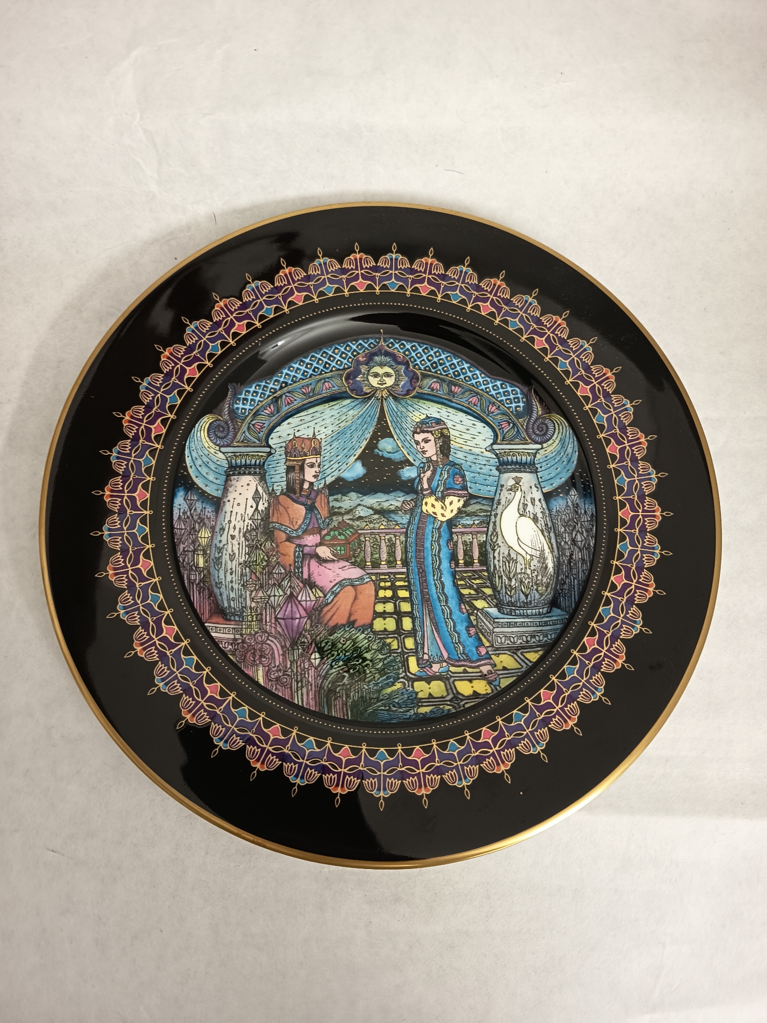 The image shows a decorative plate with a black background and a multicolored, ornate border. The central image on the plate depicts two figures, a king and a queen, in a fantastical setting. The king is seated, wearing a crown and orange robes. The queen stands next to him, wearing a blue dress and holding a yellow object. They are on a terrace with a patterned floor, under an archway adorned with a sun. Columns with carved features flank them, and a cityscape is visible in the background. The scene appears to be inspired by a fairy tale or legend. The location "Wangen bei Olten, Switzerland" is not depicted, but the plate itself may originate from that area.
