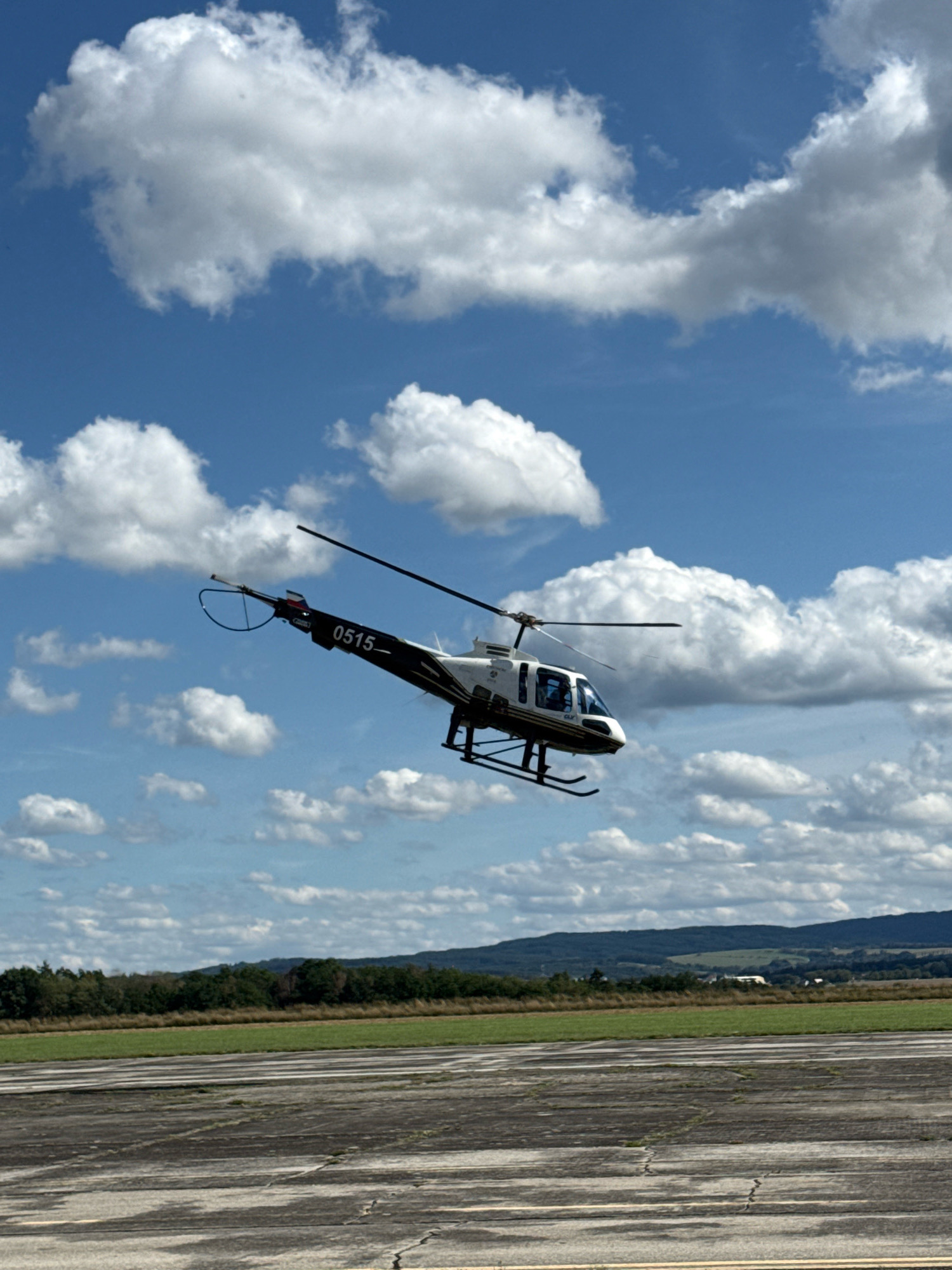 A white helicopter with black accents and the number "0515" on its tail is in mid-air against a blue sky with scattered white clouds. The helicopter is angled to the right, with its rotor blades visible. Below the helicopter is a landscape with green fields, trees, and distant hills. In the foreground, there is a concrete surface that appears to be a runway or helipad. The scene is likely in or near the city of Cheb, Czechia, given the provided context.