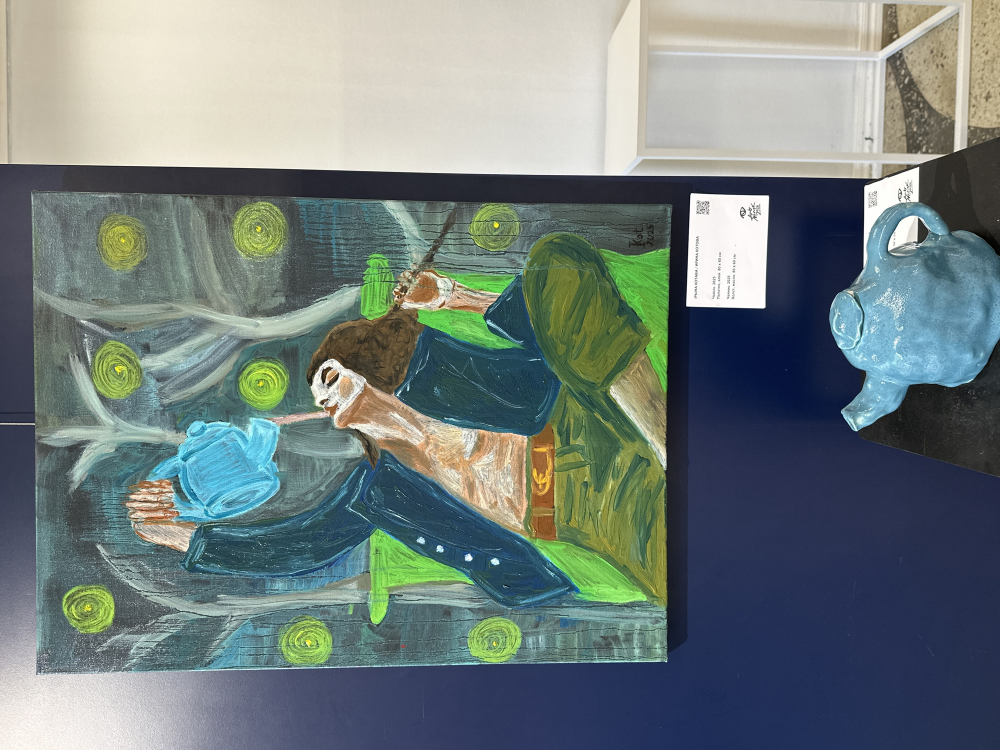 The image captures an art exhibition in Minsk, Belarus, featuring a painting and a ceramic sculpture displayed on a dark blue wall.
The central subject is an oil painting, oriented horizontally, depicting a stylized human figure with long brown hair, fair skin, and a white, mask-like facial area around the mouth and nose. The figure wears a dark blue jacket/shirt with white buttons and green trousers, cinched with a brown belt, and is shown holding and tilting a light blue watering can or teapot. The background is composed of dark, swirling blues and greens, punctuated by several luminous, concentric green-yellow circles, creating a mystical or dreamlike atmosphere reminiscent of Vincent van Gogh's style. An inscription "Kot 2015" is visible in the upper right corner of the painting.
Adjacent to the painting, on a dark display surface, is a light blue, ceramic sculpture of a teapot or watering can, echoing the object in the painting.
An information card is attached to the wall, providing details about the artwork. The artist is identified as ІРЫНА КОТАВА / ИРИНА КОТОВА (Iryna Kotava / Irina Kotova). The title of the artwork is "Чайнік. 2025" (Teapot. 2025), and the medium is specified as "Палатно, алей. 80 х 60 см." / "Холст, масло. 80 х 60 см." (Canvas, oil. 80 x 60 cm.). The card also includes an "Art Model" logo and a QR code. The setting appears to be an indoor gallery or exhibition space with ambient lighting.