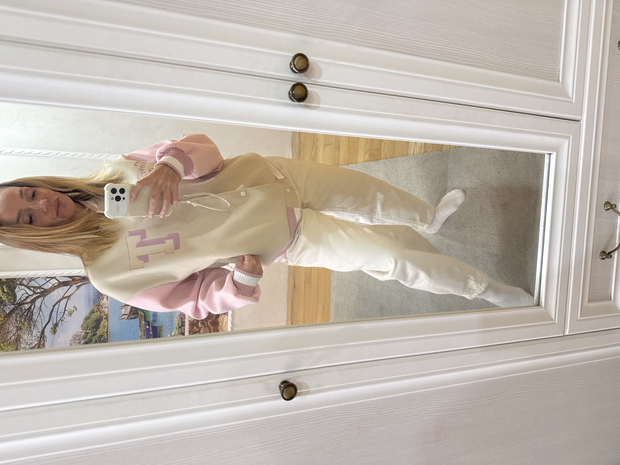 A woman is taking a selfie in a mirror. She is wearing a beige varsity jacket with pink sleeves and a purple "T" on the back, and white pants. The mirror is framed by a white cabinet with bronze knobs. The reflection in the mirror shows a glimpse of a wooden floor, a carpet, and a wall with a framed image of a coastal scene with buildings, trees, and boats. The image does not contain enough information to determine a specific location in the city of Vsevolozhsk, Russia.